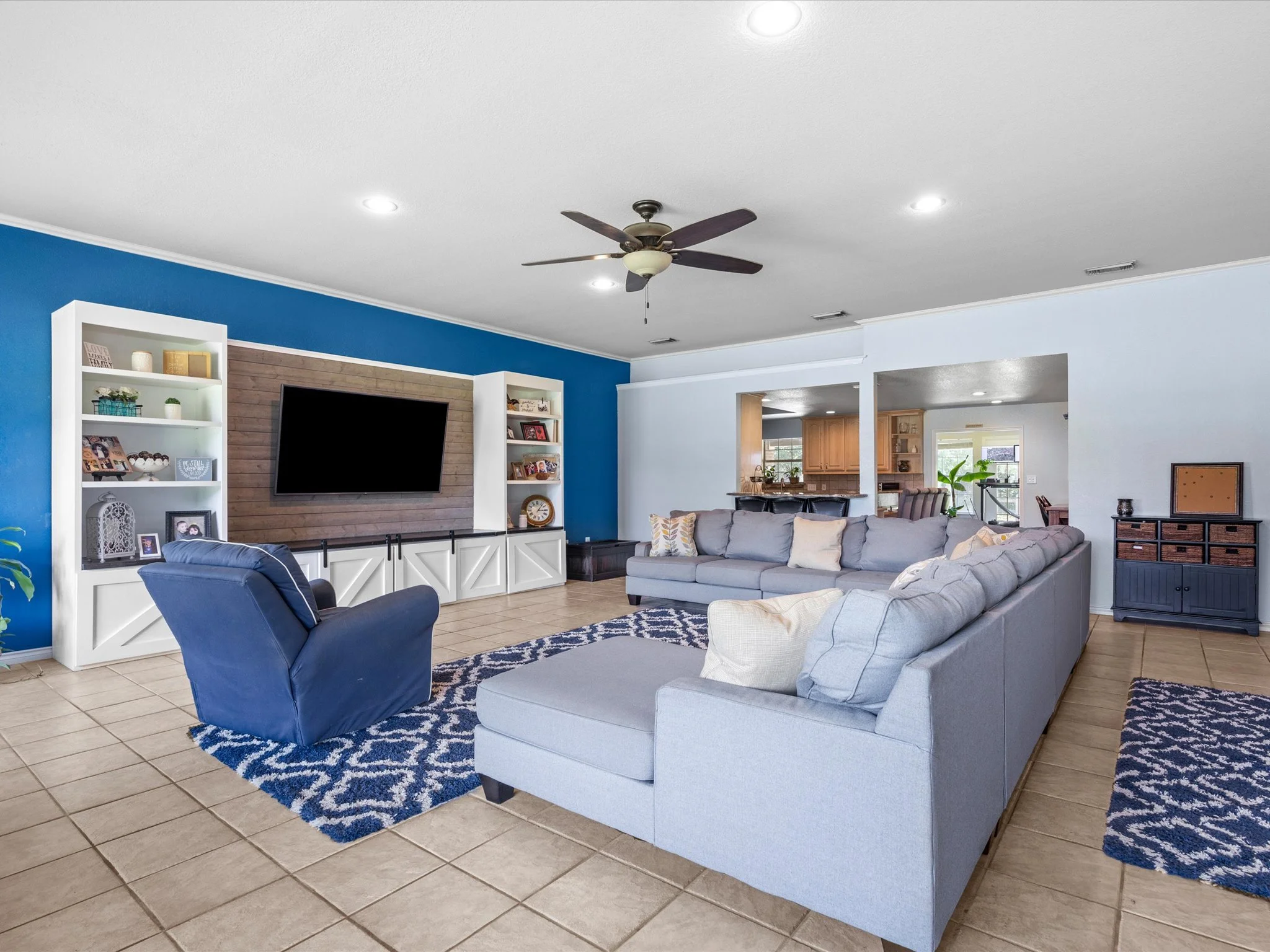 Living room with a white and blue color scheme, gray sectional sofa, blue armchair, patterned rug, and wall-mounted TV, with a ceiling fan overhead.