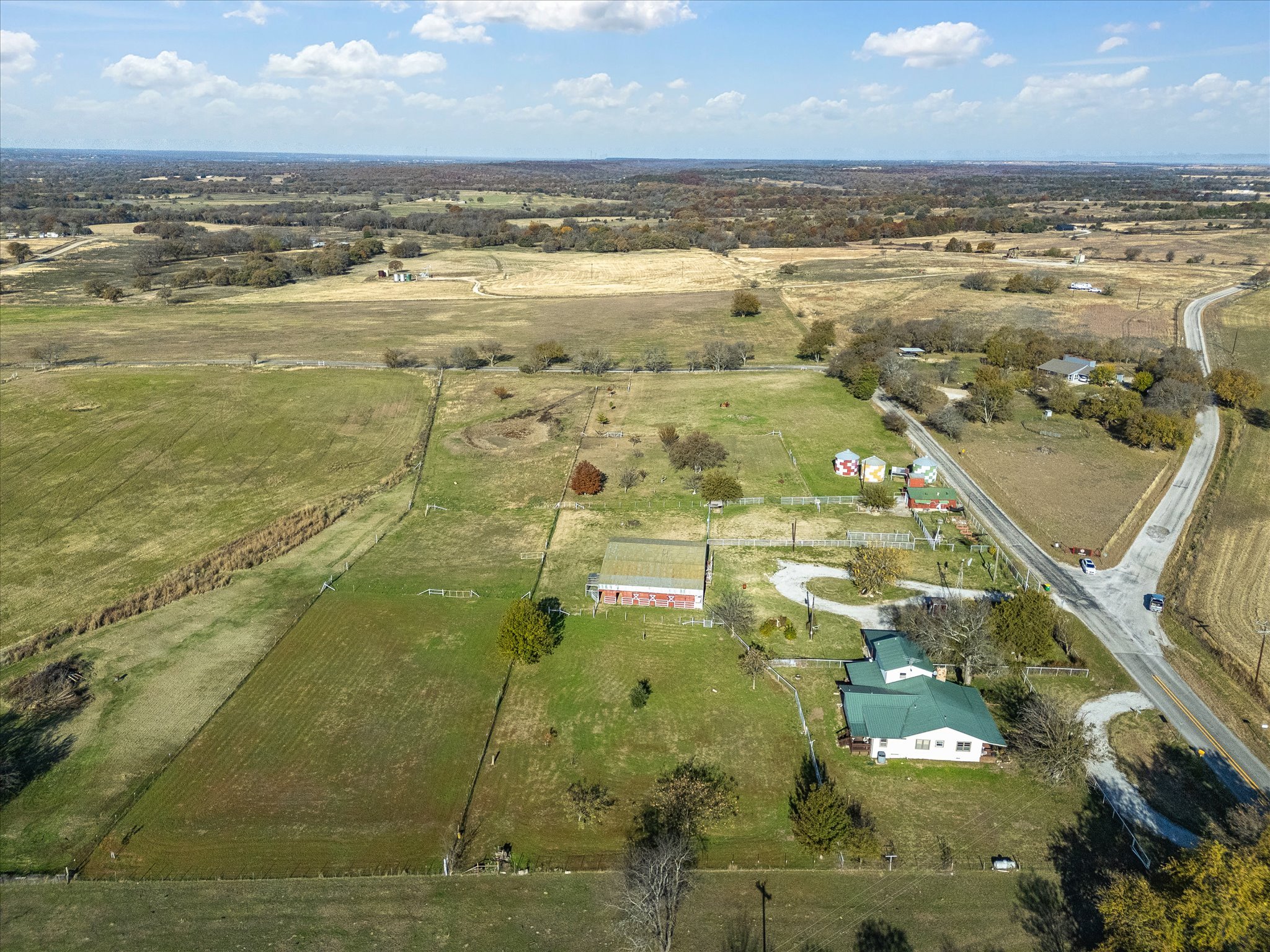 Aerial view of a rural landscape with open fields, scattered trees, houses, and winding roads under a partly cloudy sky.