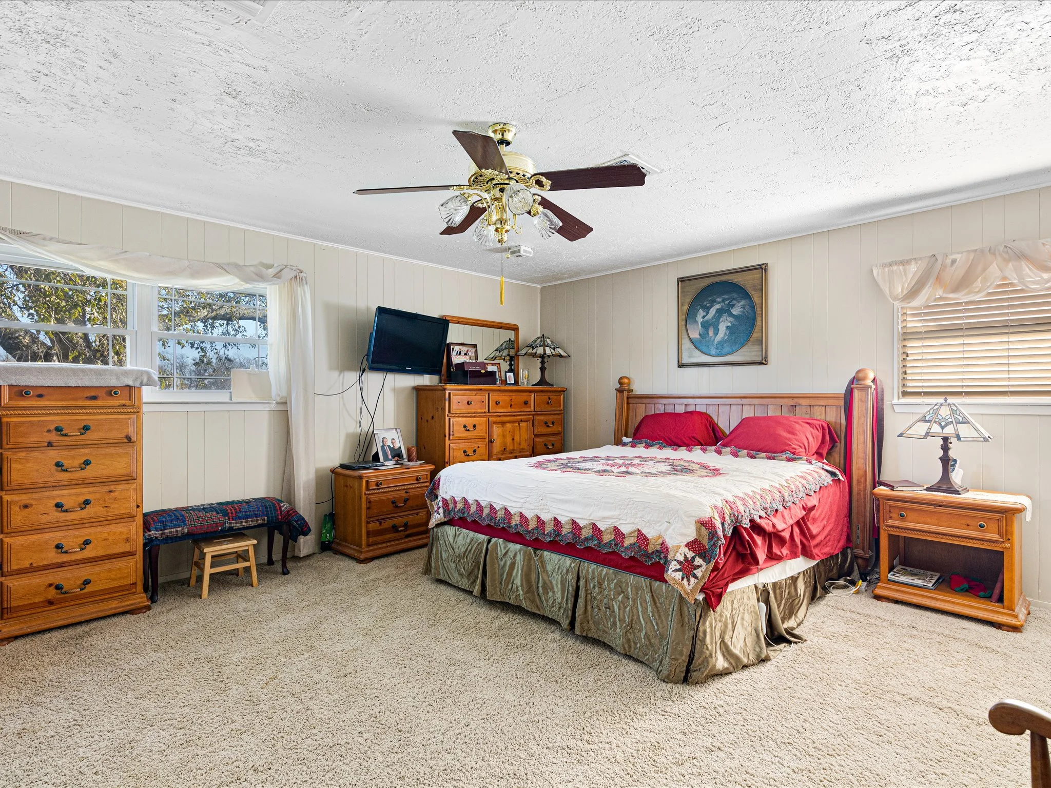 A bedroom with a bed covered in a quilt with red and white patterns, wooden dressers, a wall-mounted TV, a framed picture of a horse, and a ceiling fan with light fixtures.