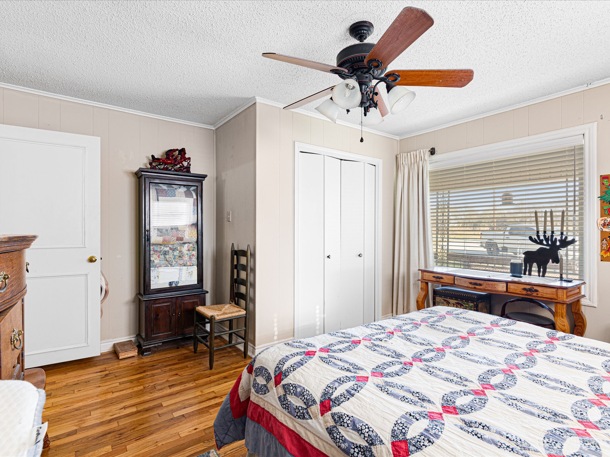 A bedroom with a ceiling fan, a quilted bedspread, a window with blinds, and a wooden dresser with a reindeer figurine.