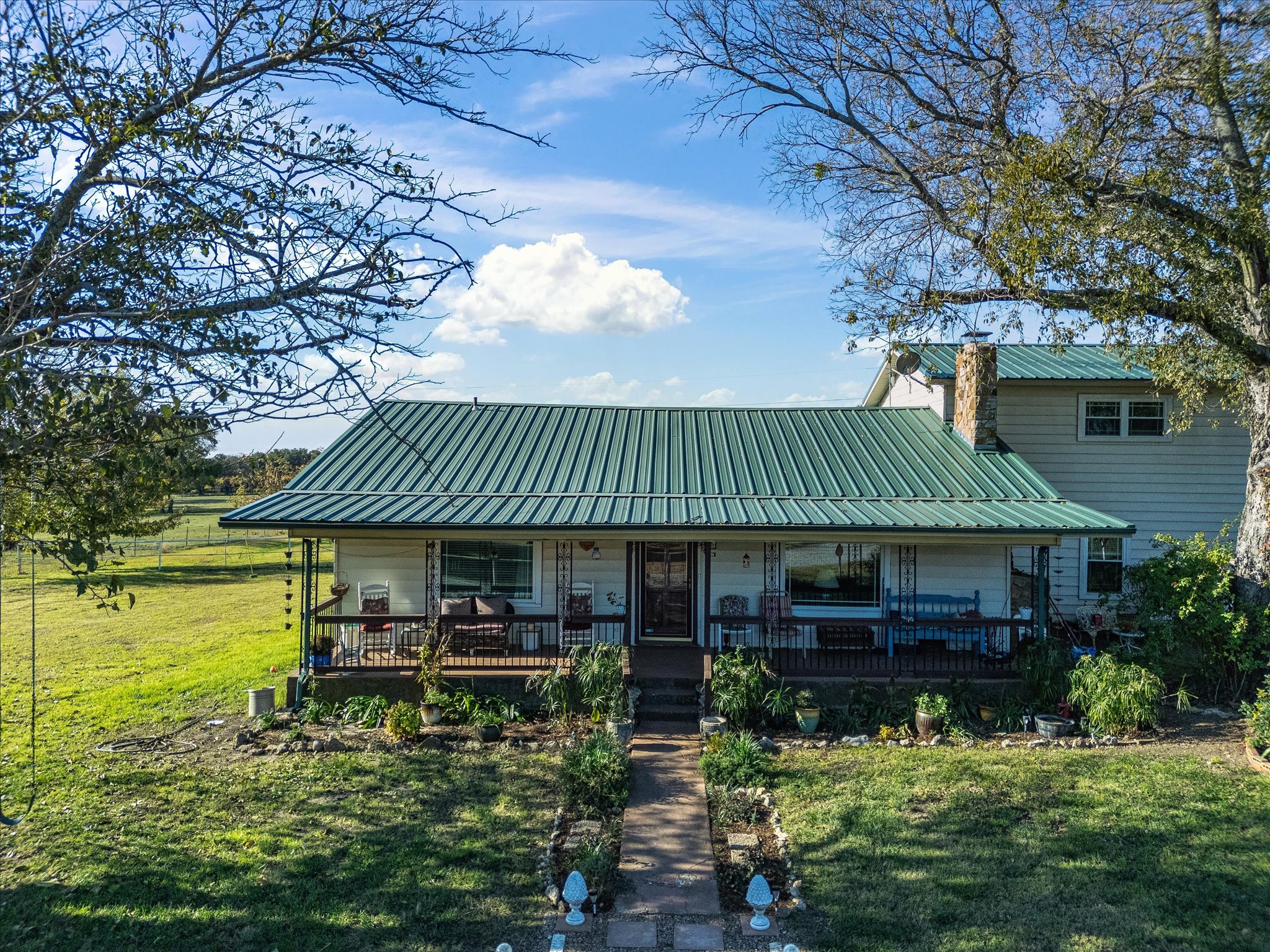 A house with a green metal roof, a front porch with chairs, and a garden with various plants. There are two large trees, one with leaves and one without, and a blue sky with scattered clouds in the background.