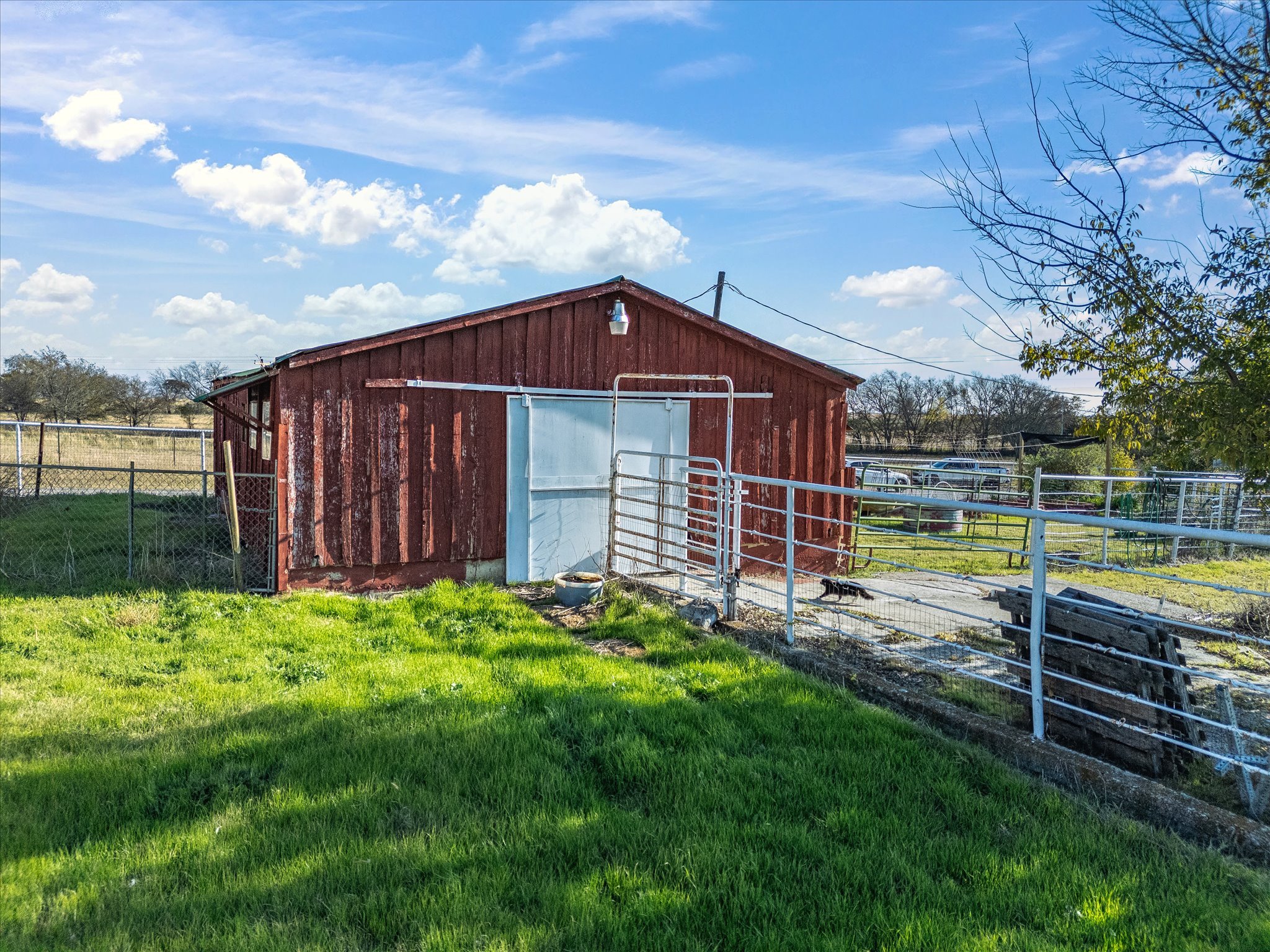 A red wooden barn with a metal door and a light fixture on the roof, surrounded by a fenced yard with green grass and a clear blue sky with white clouds.