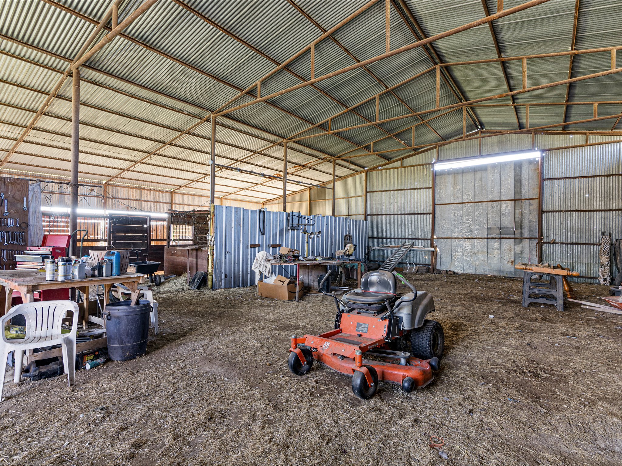 Inside a rustic workshop or barn with a metal roof and walls. The floor is dirt, and there are various tools, parts, and equipment scattered around, including a lawn mower on the ground.