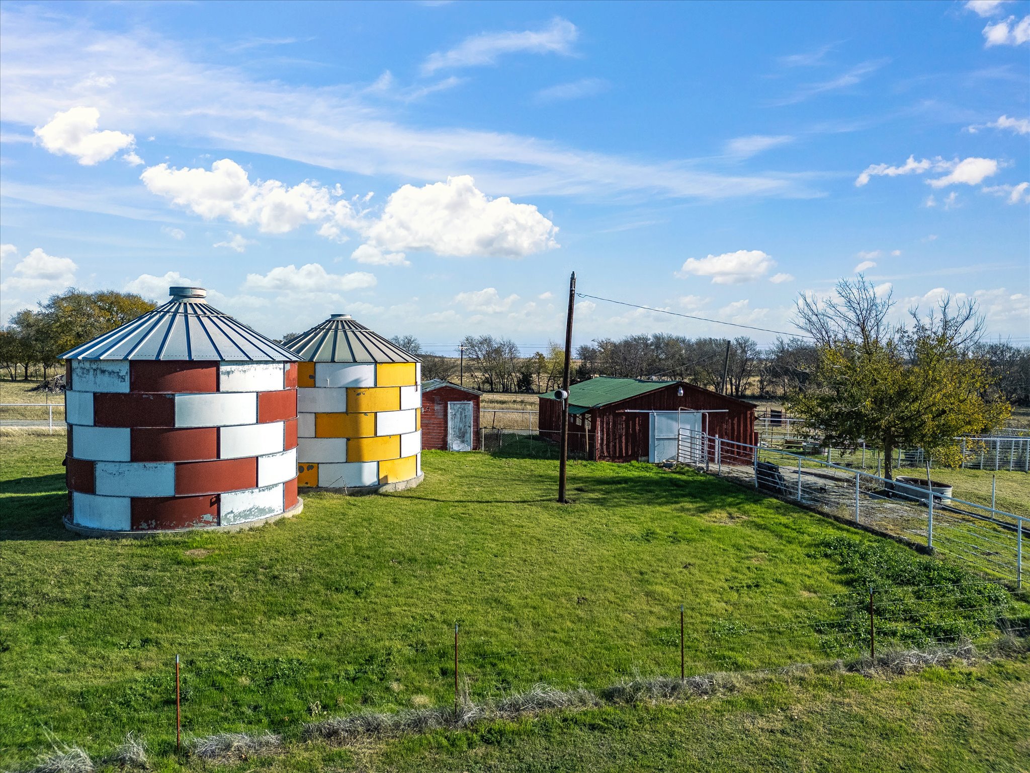 Two colorful rounded storage tanks with metal roofs, one painted with red, white, and brown stripes and the other painted with white, yellow, and green stripes, in an open grassy field under a blue sky with scattered clouds, surrounded by trees and f