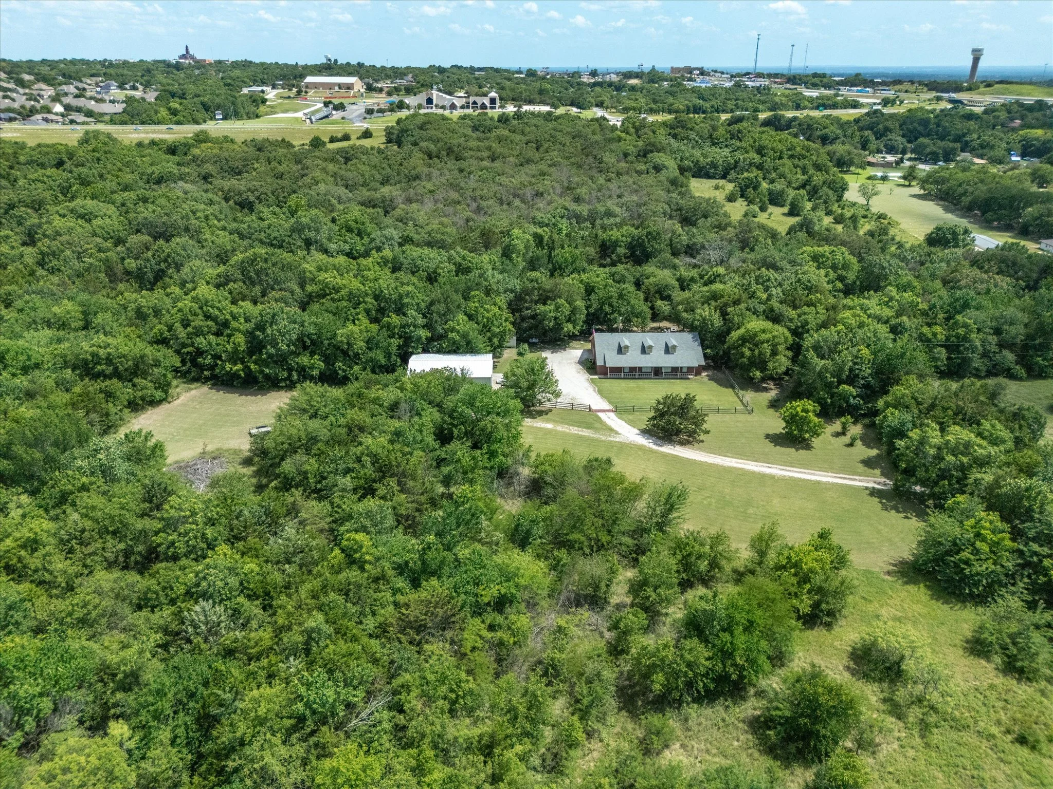 An aerial view of a lush, green forest with a small house in the lower part of the image, and a commercial area and highways in the background under a blue sky with some clouds.