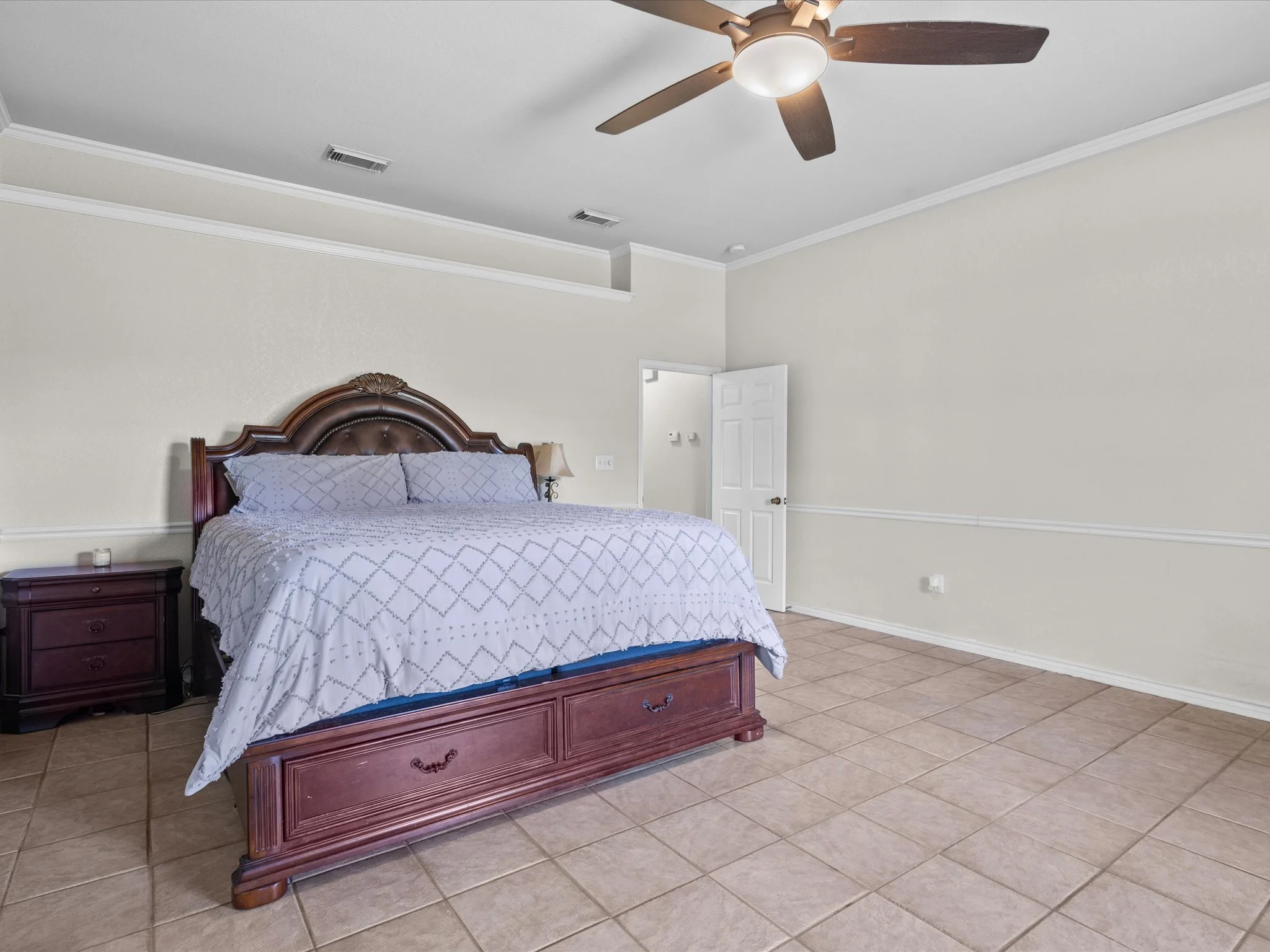 A bedroom with a wooden bed frame, gray and white bedding, a nightstand with a lamp, a ceiling fan with wooden blades, and tile flooring, with a partially open door and white walls.