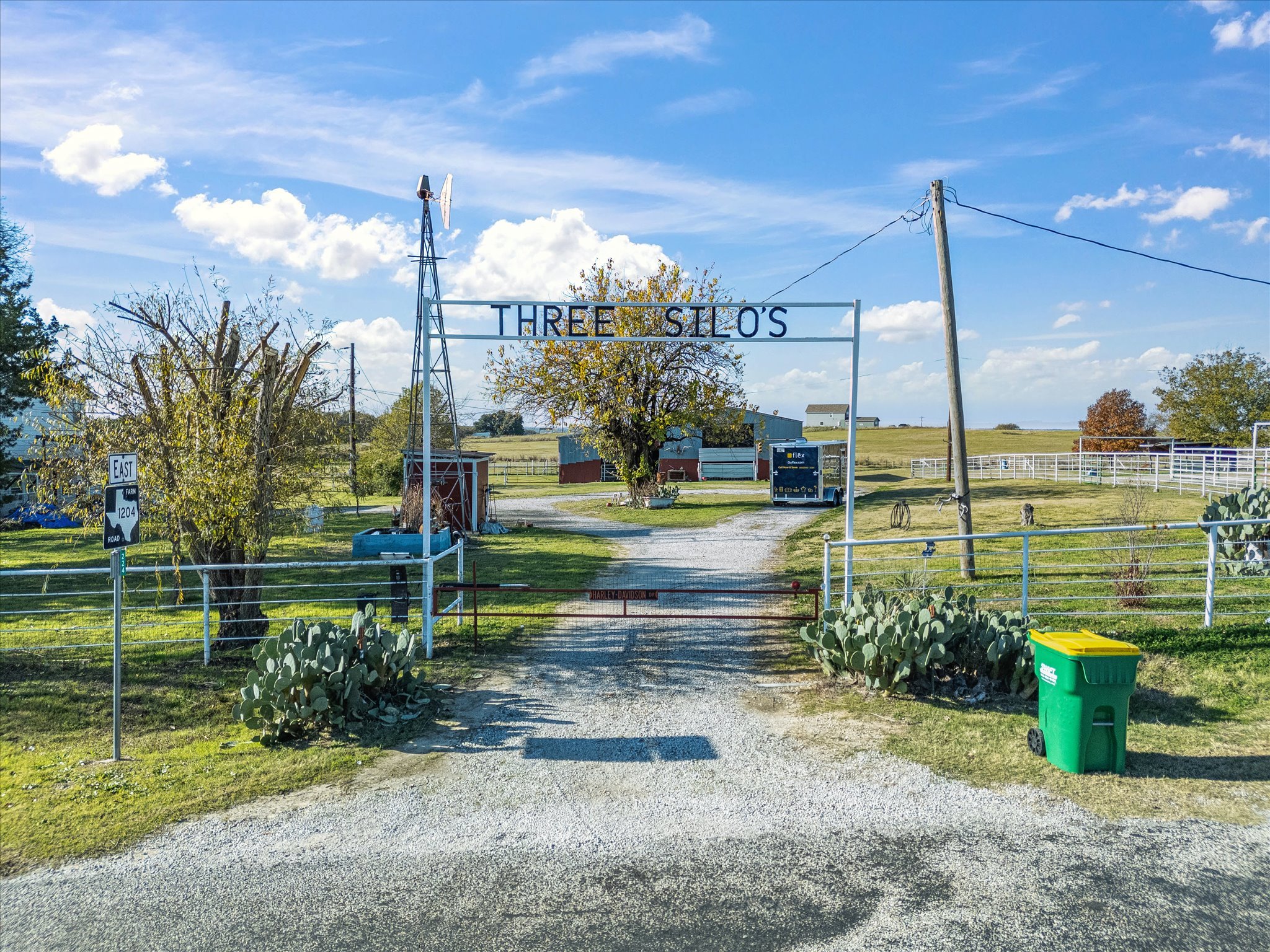 A rural scene with a gravel driveway leading to a farm, featuring a metal sign that reads 'THREE SILO'S'. There are trees, farm buildings, and a power pole with wires against a blue sky with scattered clouds.