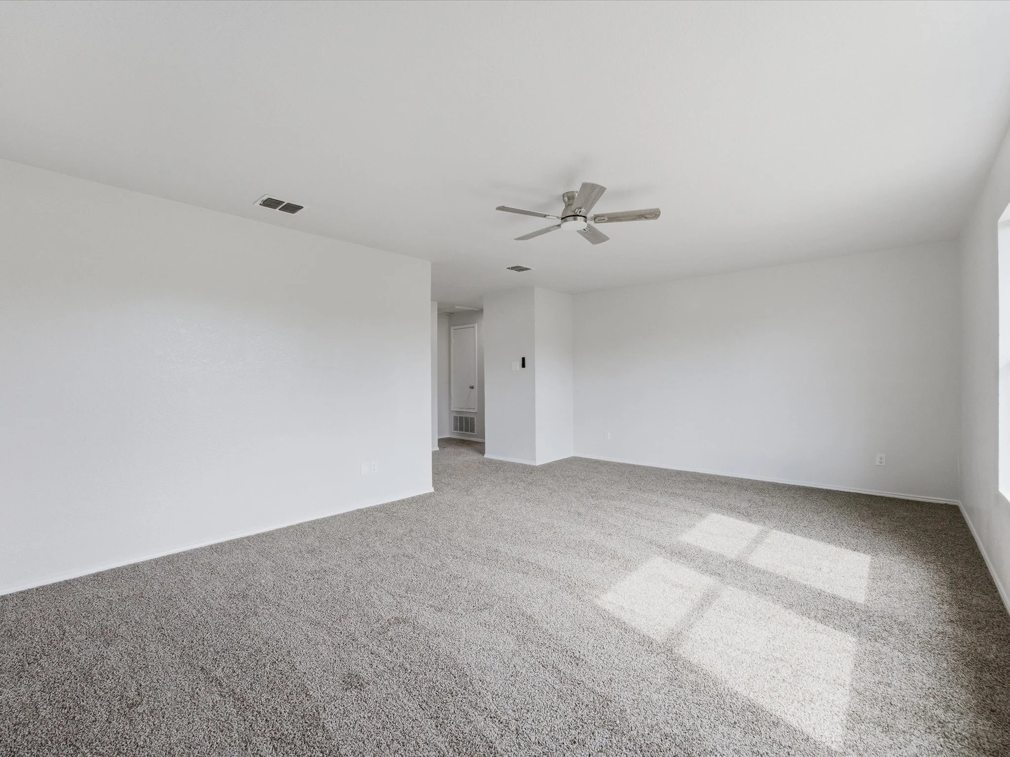 Empty living room with white walls, beige carpet, ceiling fan, and vents.