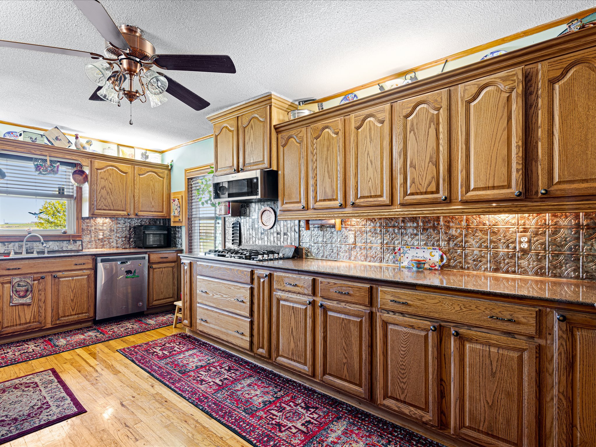 Kitchen with wooden cabinets, a microwave, a dishwasher, and decorative plates on top. There is a window with blinds, a ceiling fan with lights, and a patterned rug on a wooden floor.
