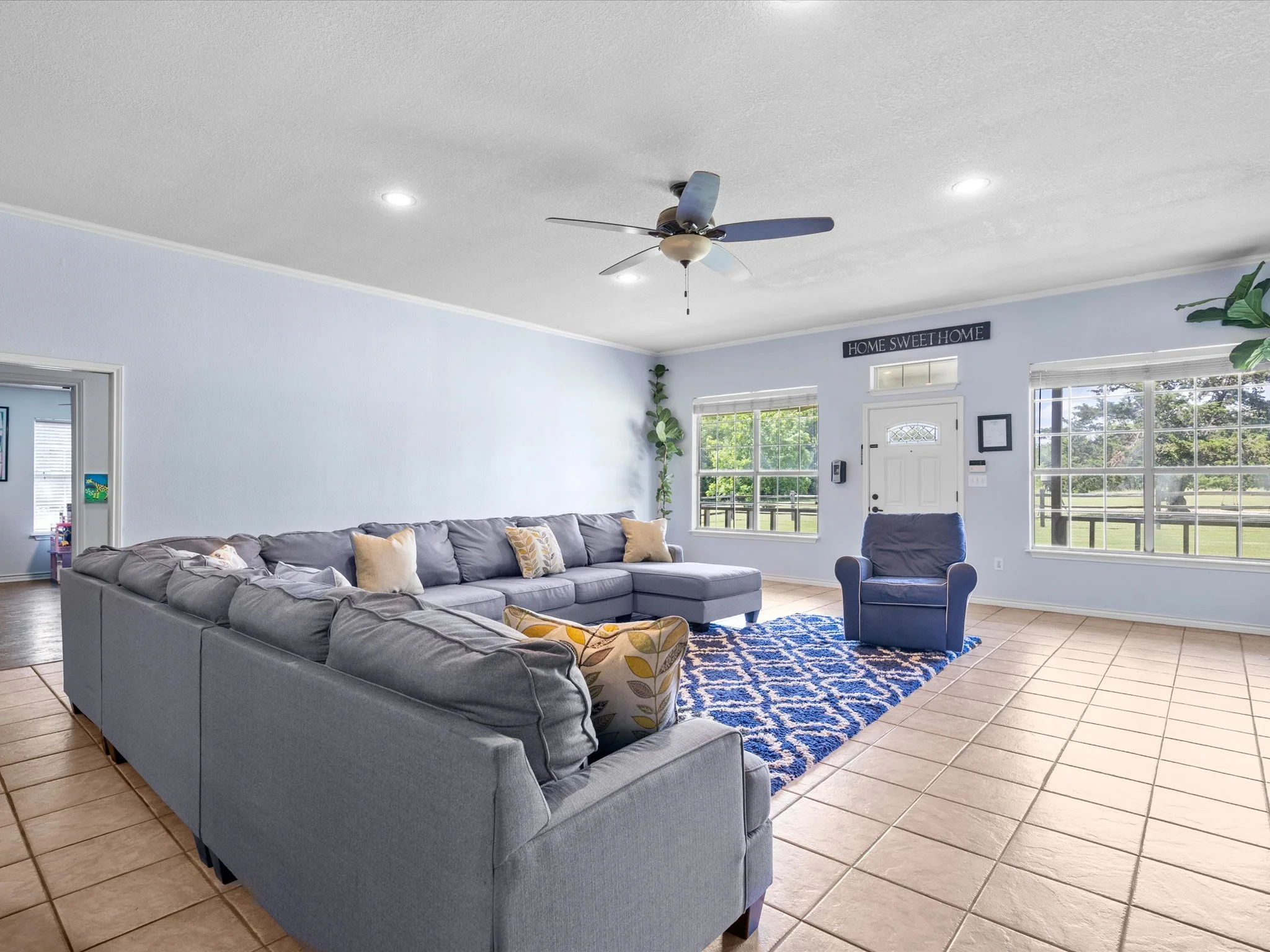 Interior of a living room with a large gray sectional sofa, decorative pillows, a blue patterned rug, and a blue armchair. The room has light blue walls, large windows allowing natural light, and a ceiling fan. A sign above the door reads "Home Sweet