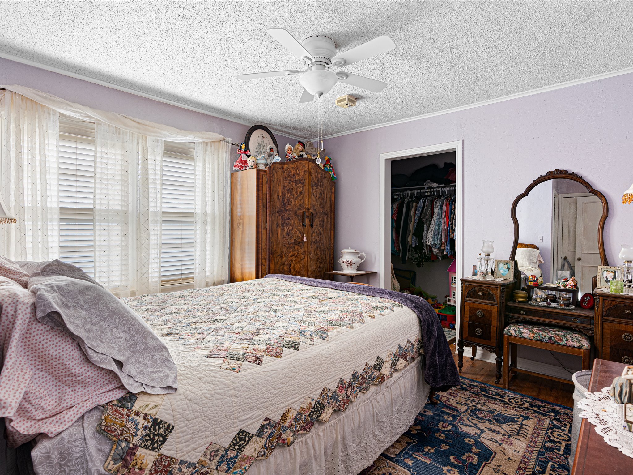 A bedroom with purple walls, a ceiling fan, a dresser with dolls on top, a closet with clothes, a mirror, and a bed with a quilt and pillows.
