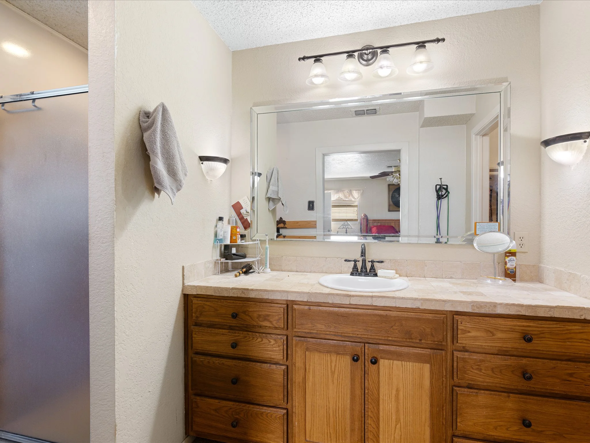 Bathroom vanity with a large mirror, a wooden cabinet with drawers, and a beige countertop with a sink. Various toiletries and grooming items are on the counter. There are two wall-mounted light fixtures above the mirror and a towel hanging on the wa