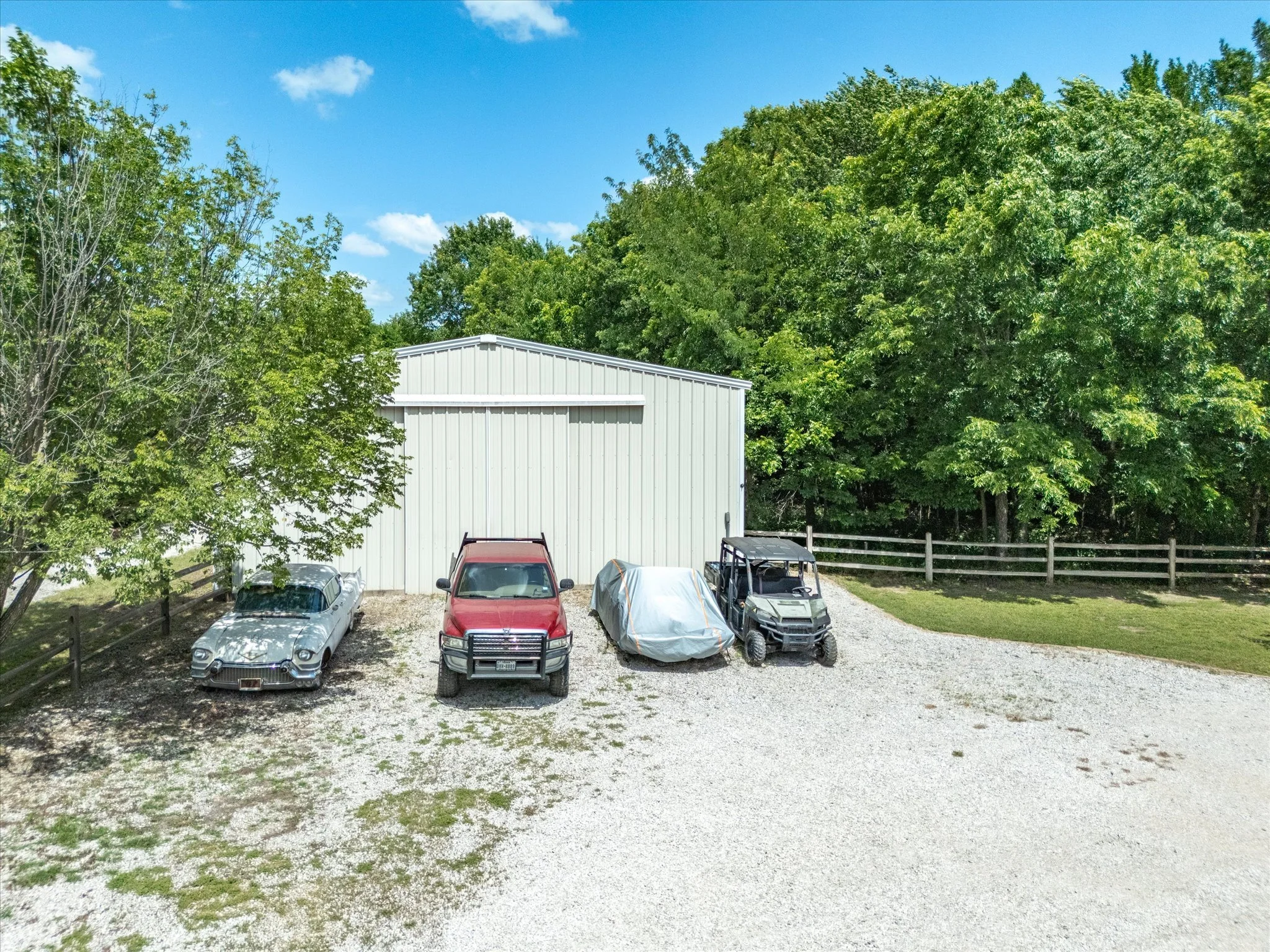 A gravel driveway with four vehicles parked in front of a white metal building, surrounded by green trees and a blue sky with scattered clouds.