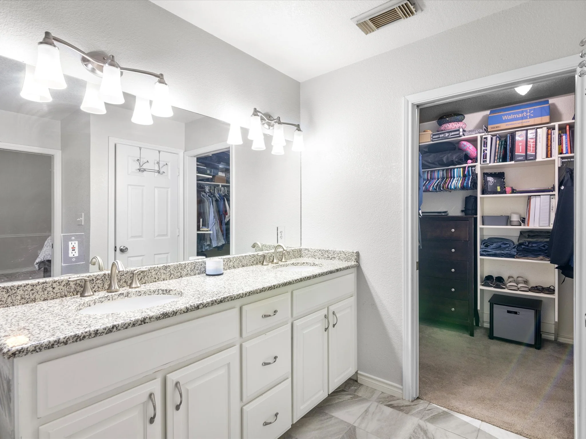 Bathroom vanity with a granite countertop and white cabinets, large mirror, overhead light fixture, open closet with clothes and storage boxes, and a doorway with a dark dresser inside.