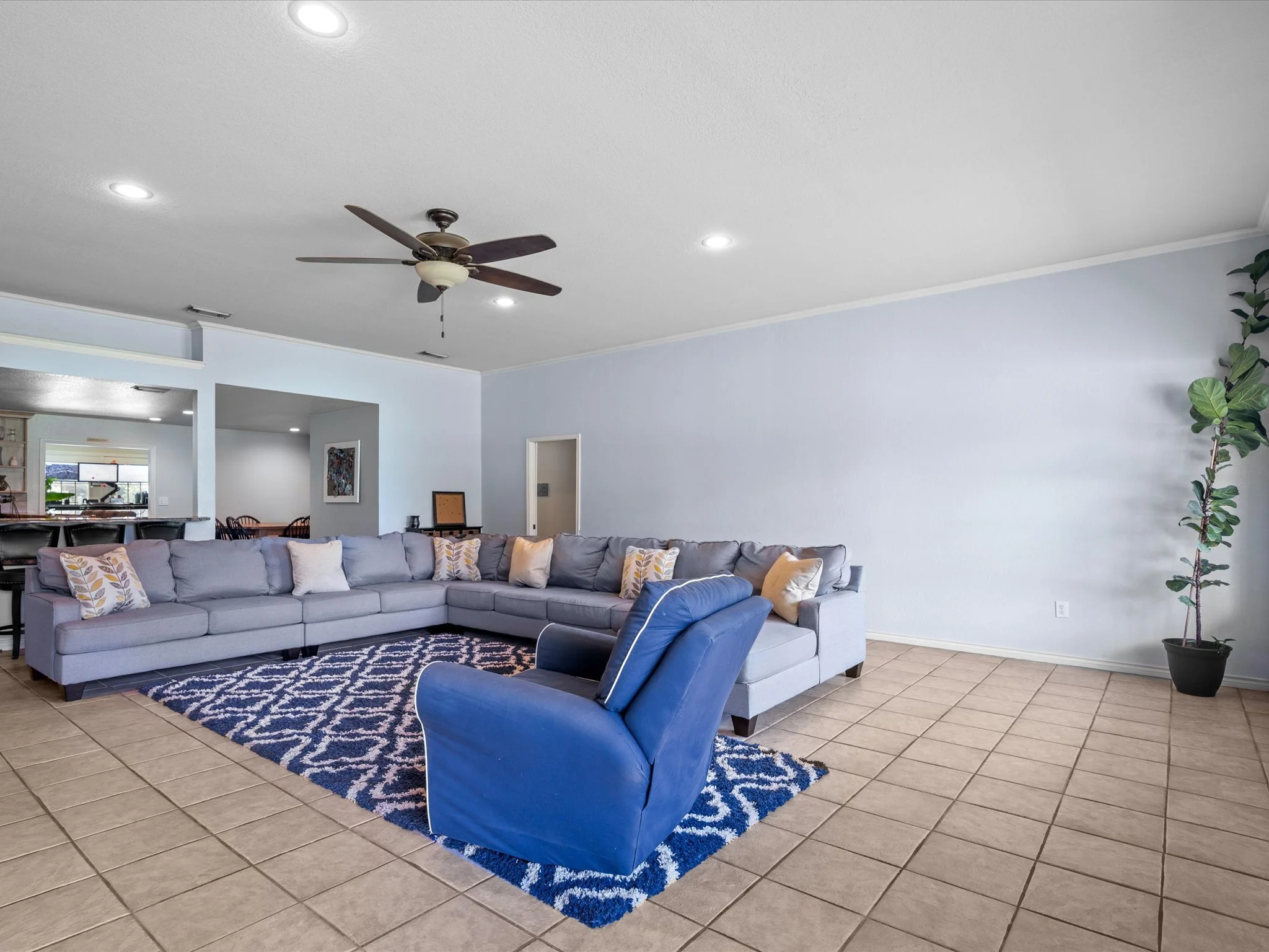 Living room with a large gray sectional sofa, a blue armchair, a patterned blue and white rug, a ceiling fan with lights, a plant in a pot, and a dining area in the background.
