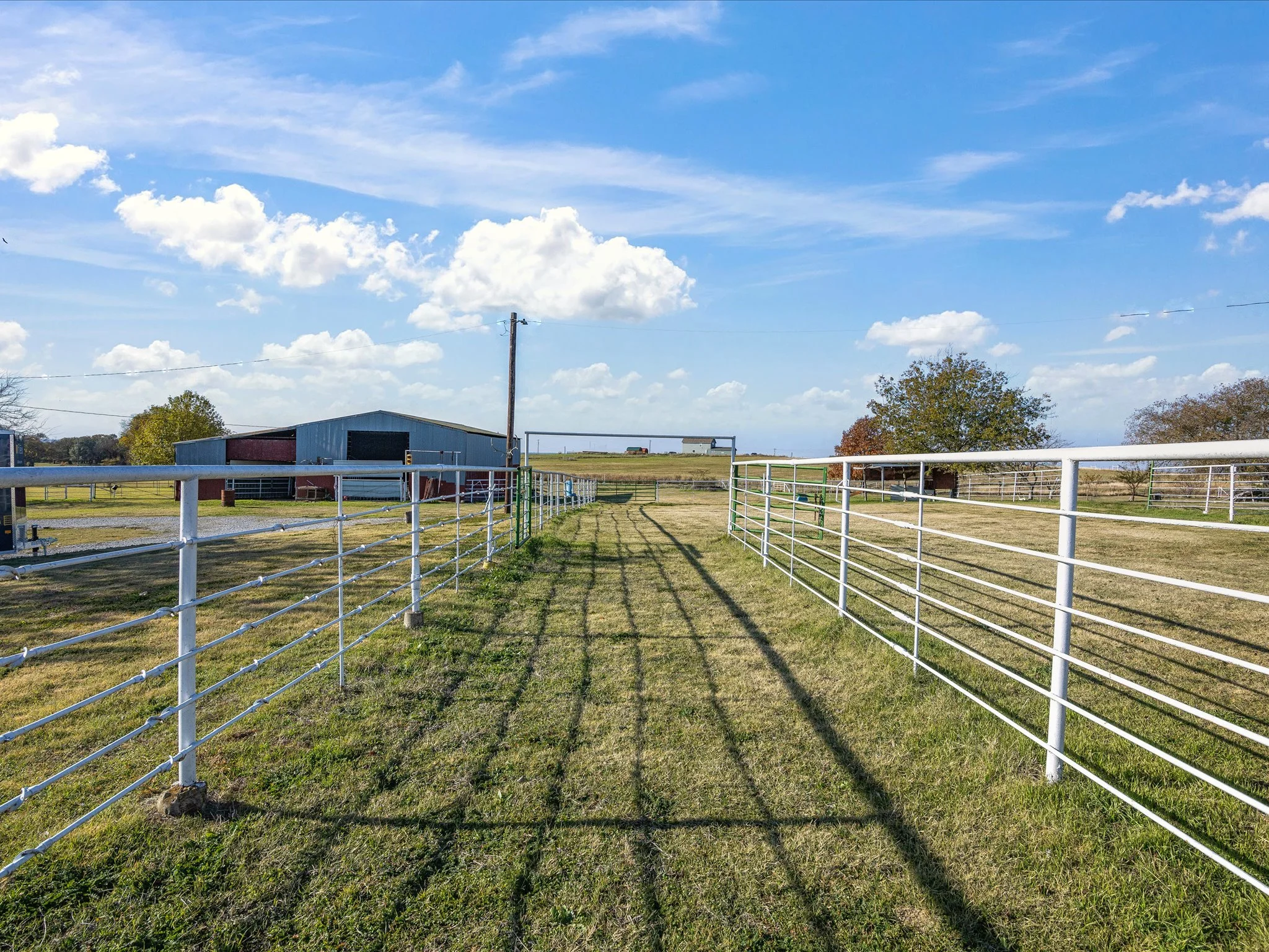 A large open field with green grass enclosed by white metal fencing, a blue barn, power lines, trees, and a cloudy sky.