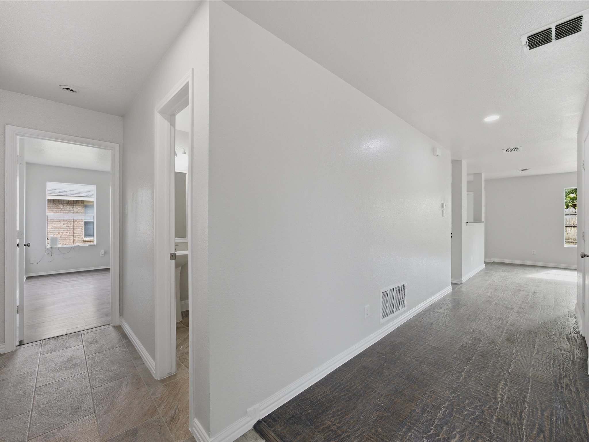 Interior view of a modern house with white walls, gray tiled and dark wooden flooring, and multiple windows allowing natural light to enter.