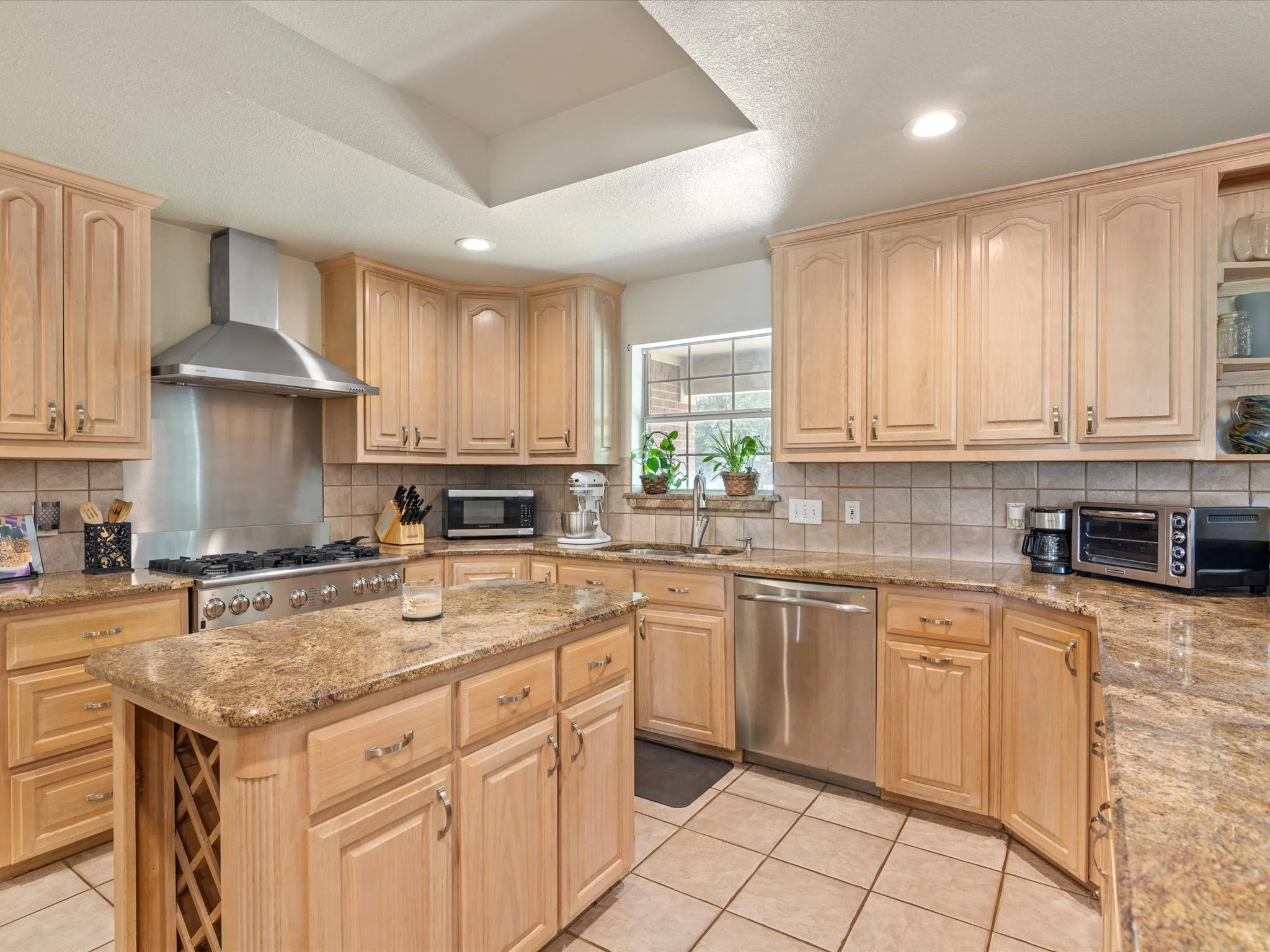 A kitchen with light wood cabinets, a granite countertop, a stainless steel stove and range hood, a microwave, a coffee maker, and a window with plants on the sill. There is a small island with a granite top in the foreground.