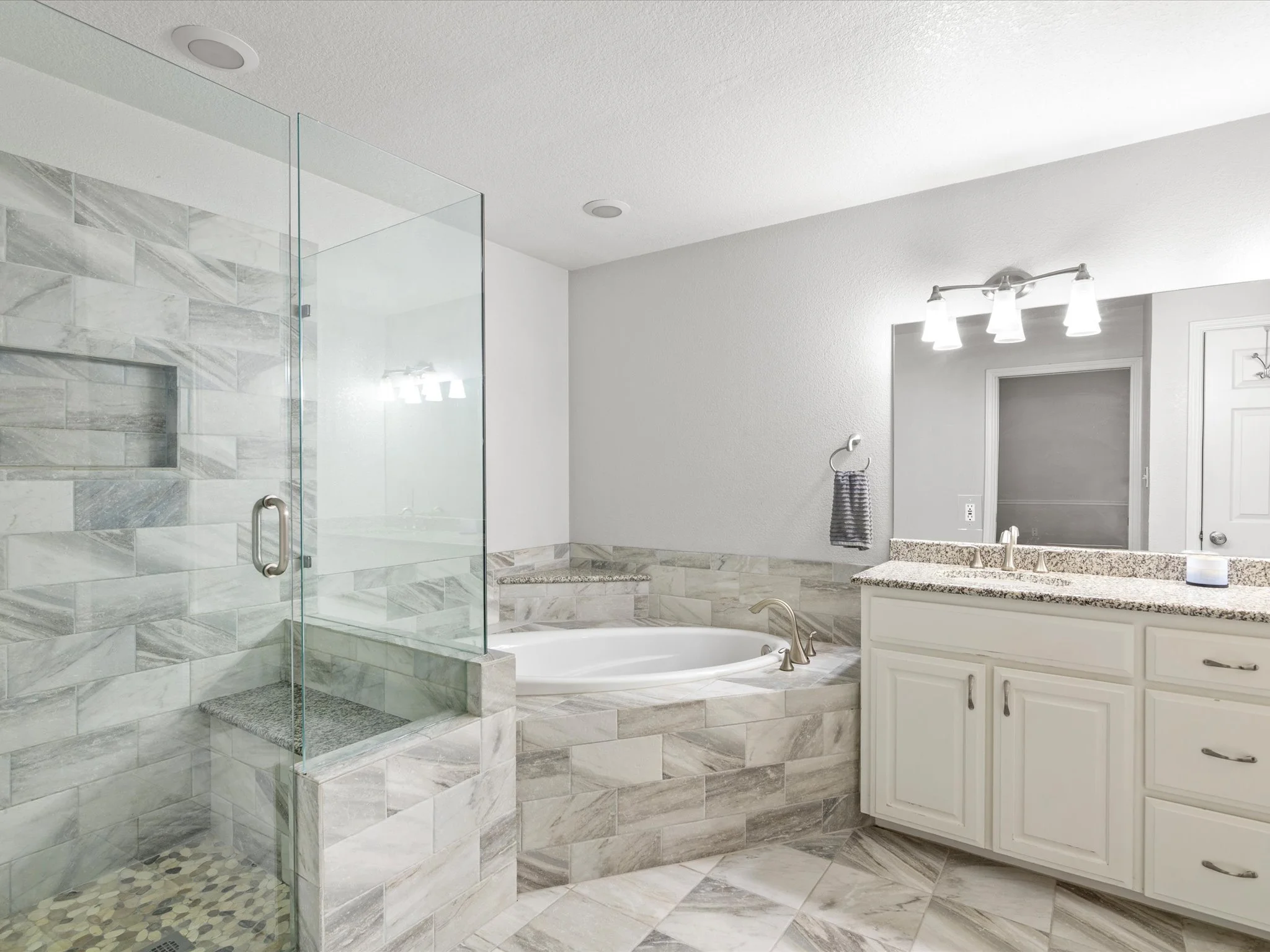 A modern bathroom with a glass shower, a bathtub, a white vanity with a granite countertop, and a large mirror with lighting fixtures above it.