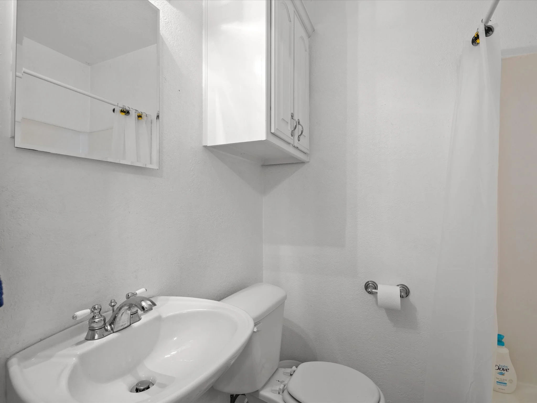 A small white bathroom with a single sink vanity, a mirror, a cabinet with doors, a toilet, and a towel holder with a roll of toilet paper. There is a bottle of baby lotion on a shelf near the toilet.