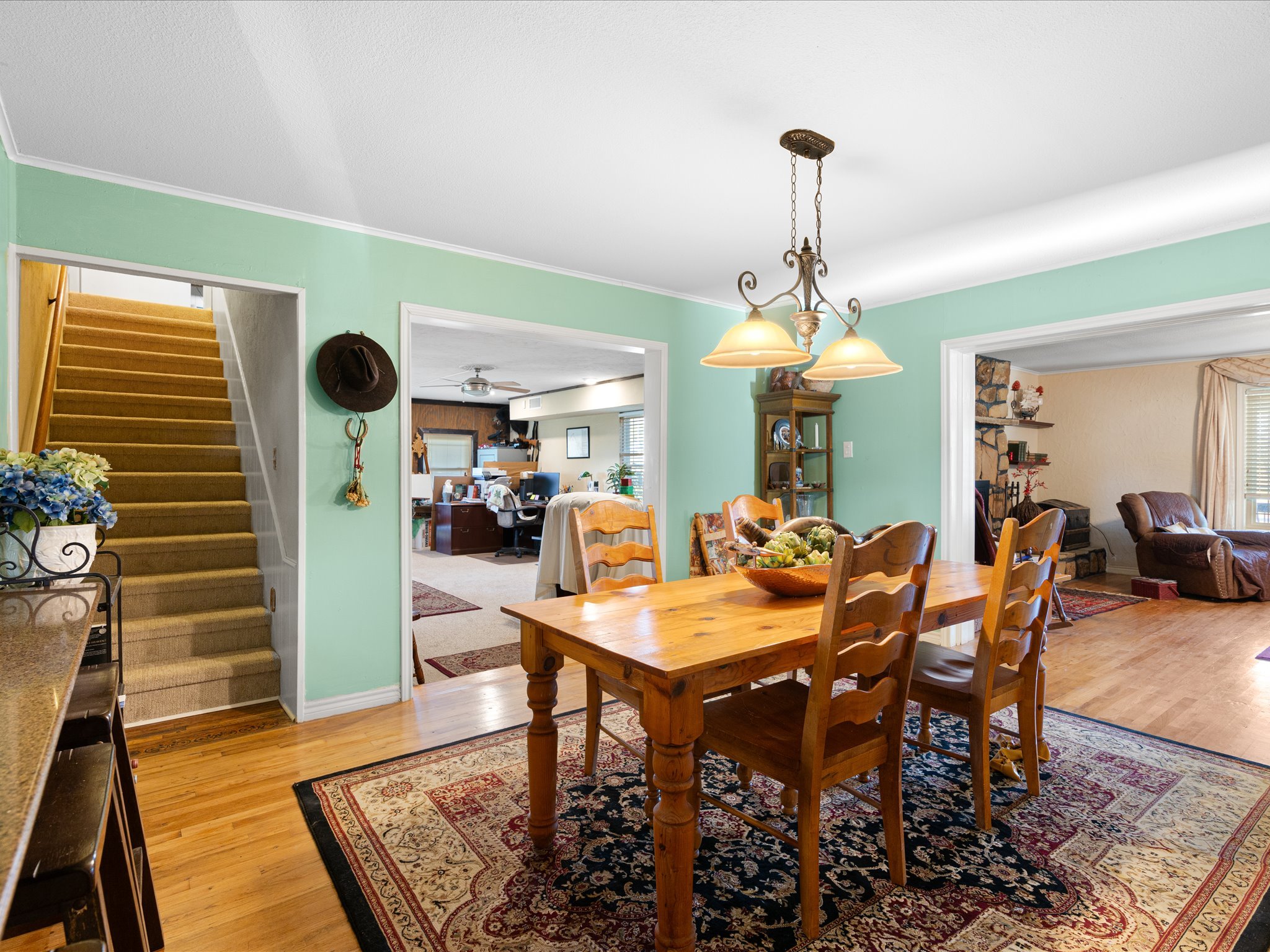 A spacious dining area and living room with a wooden dining table and chairs, a patterned area rug, a hanging light fixture, a bookshelf, and a view into a cozy living space with a stone fireplace and seating.