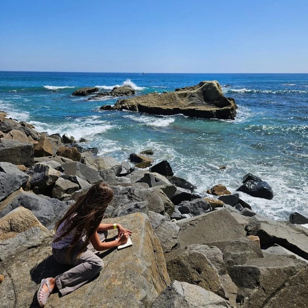 A girl with long hair is sitting on large rocks by the ocean, writing in a notebook, with the blue sea and clear sky in the background.