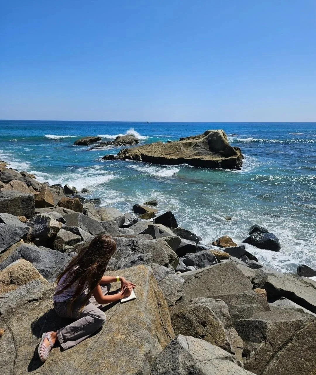 A young girl with long hair sitting on large rocks at the coastline, writing in a notebook with the ocean and waves crashing against rocks in the background under a clear blue sky.