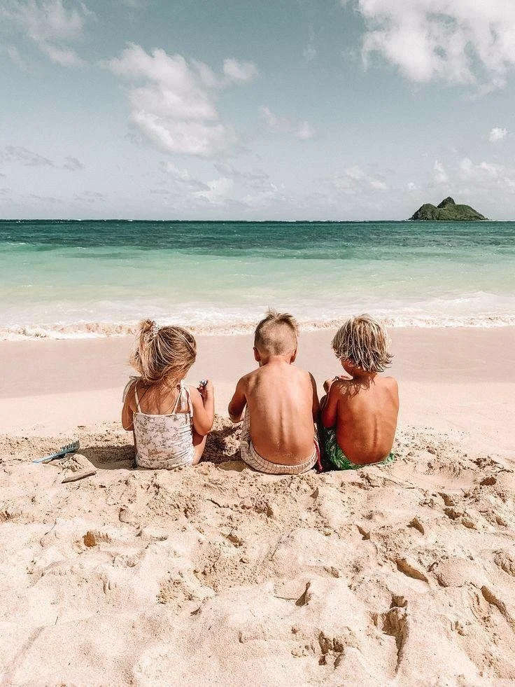 Three children sitting on a sandy beach facing the ocean, with a small island in the distance.
