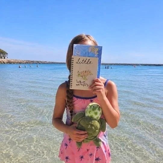 Girl at beach holding a stuffed turtle toy and a wood-burned notebook in front of her face at the beach.