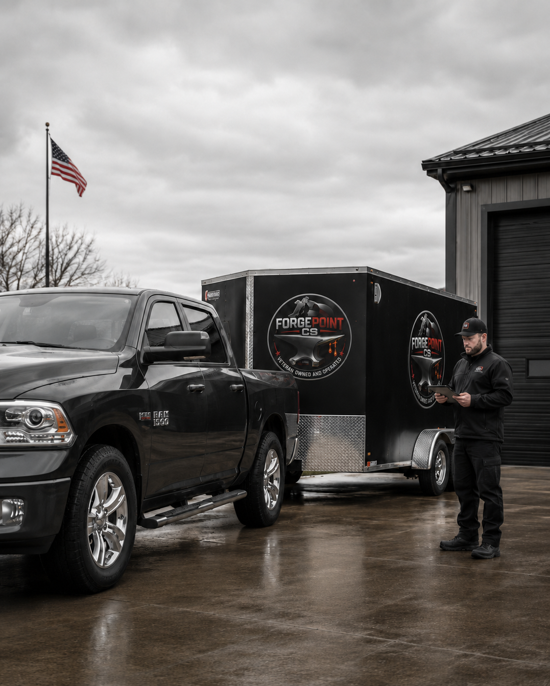 A black pickup truck towing a black trailer with 'Forge Point CS' logos parked outside on a cloudy day. A man in black clothing reads from a tablet near the trailer, with an American flag flying in the background.