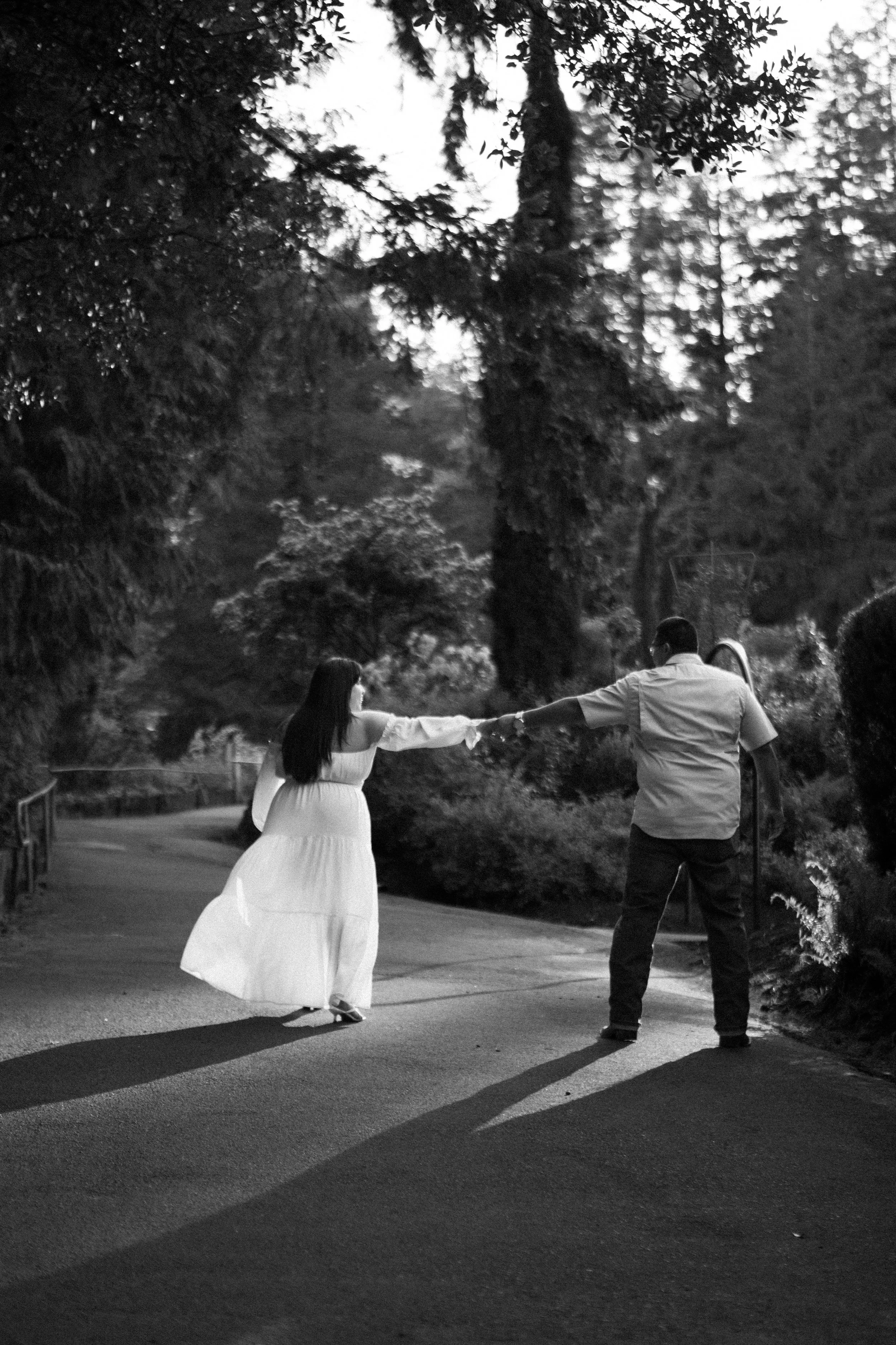 A couple holding hands and dancing on a paved path surrounded by trees in a park at sunset, in black and white.