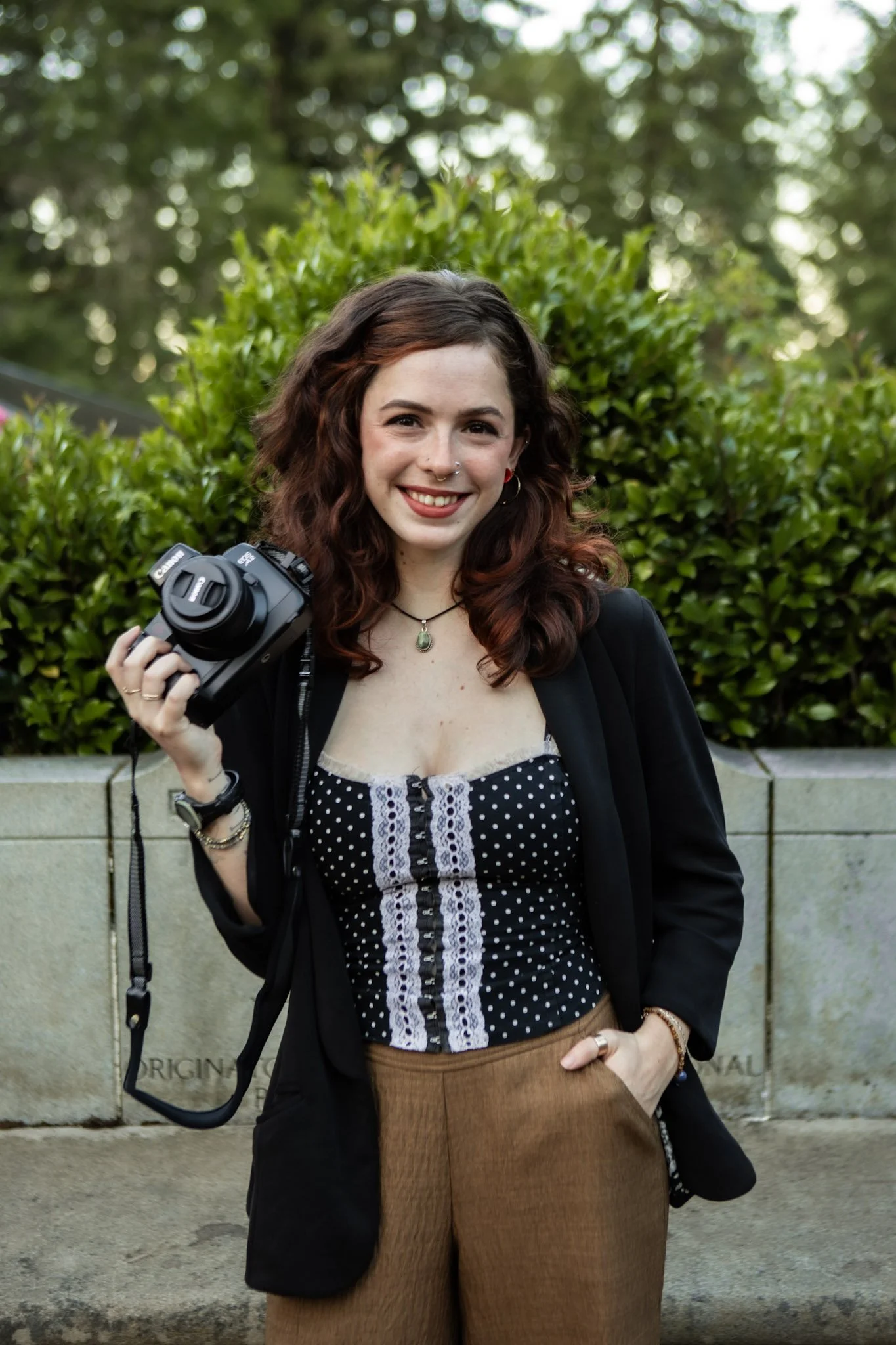 A young woman with curly brown hair holding a camera, smiling, outdoors with green foliage and a stone wall behind her.
