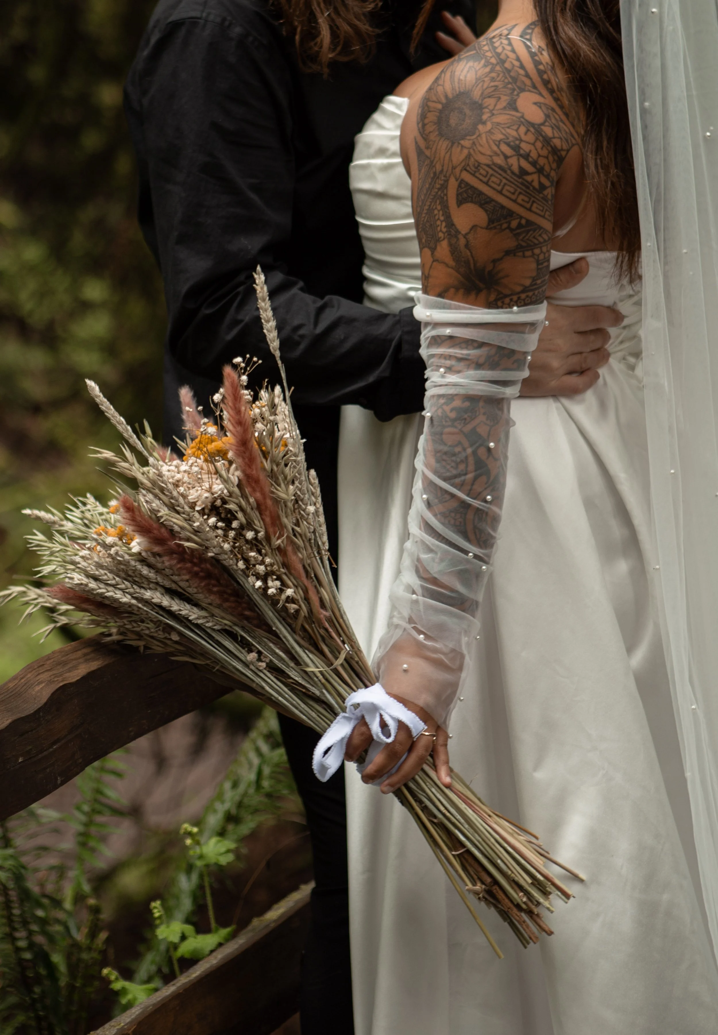A bride holding a bouquet of dried flowers and grasses, standing next to a groom at their wedding ceremony outdoors.