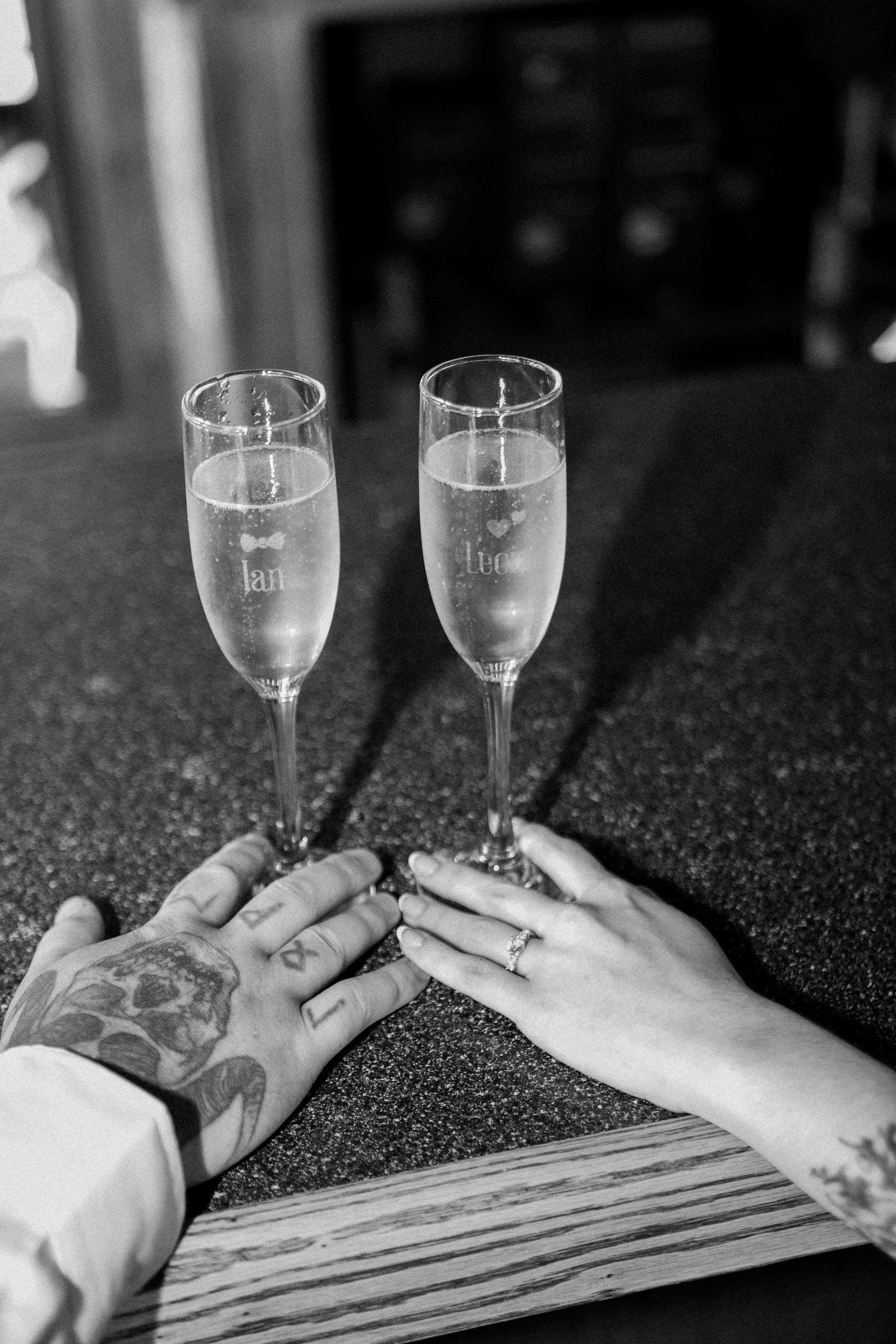 Two champagne flutes filled with sparkling wine are held by two hands, one with tattoos and a wedding ring, on a wooden table. The background is blurred.