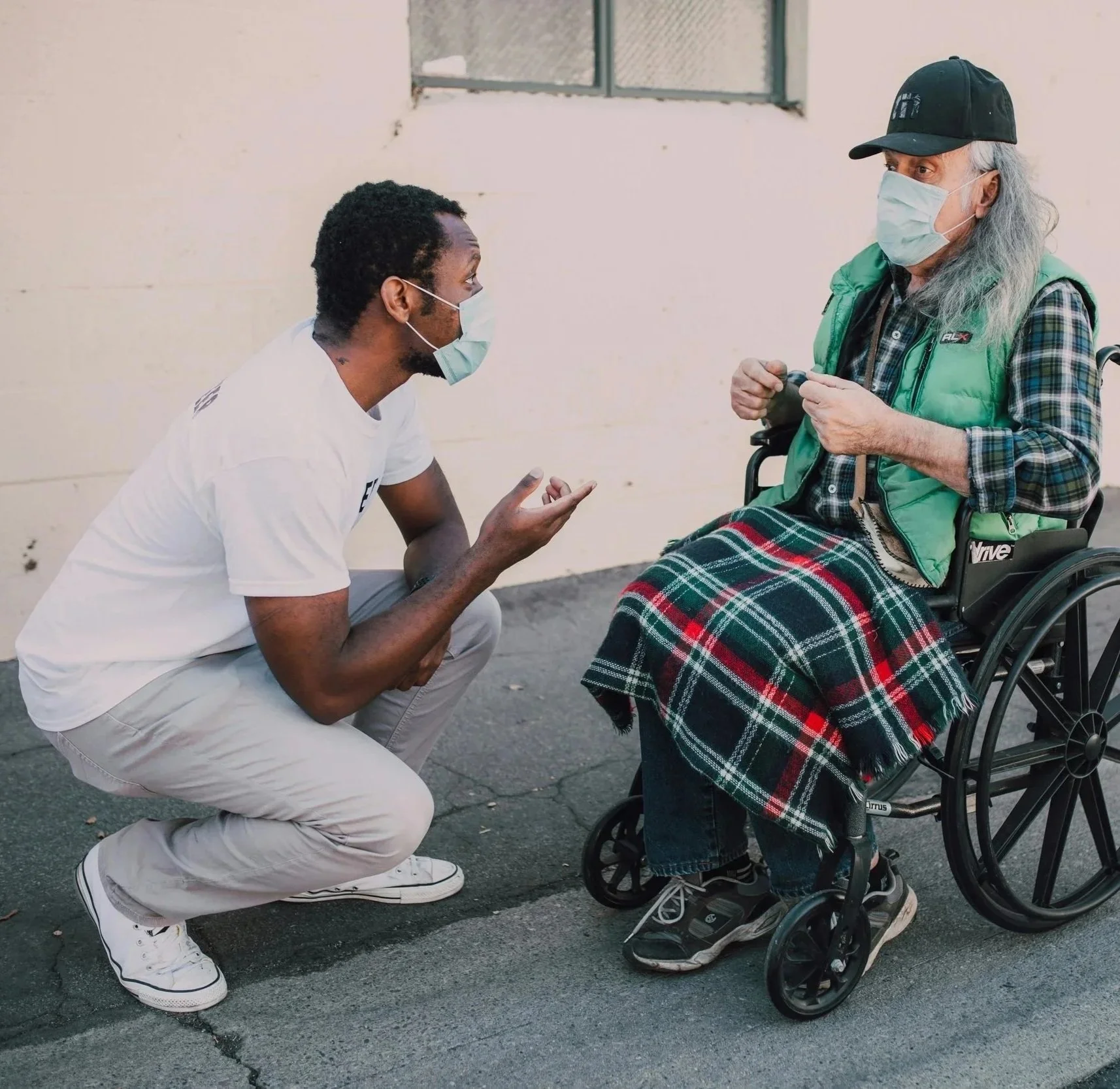 A young man kneeling and talking to an elderly woman in a wheelchair outside on a paved surface. Both are wearing face masks. The woman has long gray hair, a hat, a green vest over plaid shirt, and a plaid blanket over her lap.