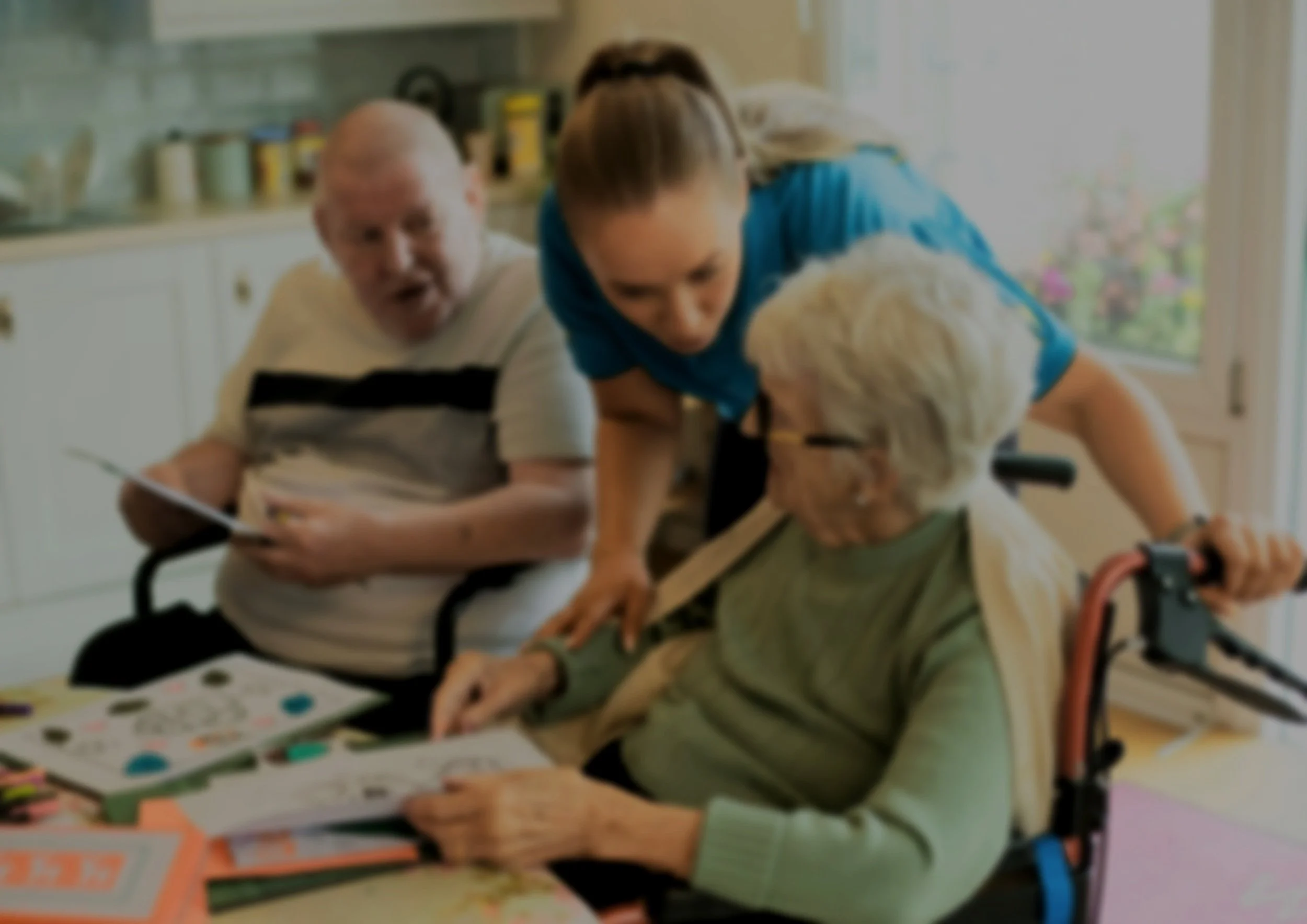 Two elderly men, one with glasses, sitting at a table with coloring books and stickers, engaging with a woman who appears to be assisting them. The setting looks like a care home or community center, with a window and shelves in the background.