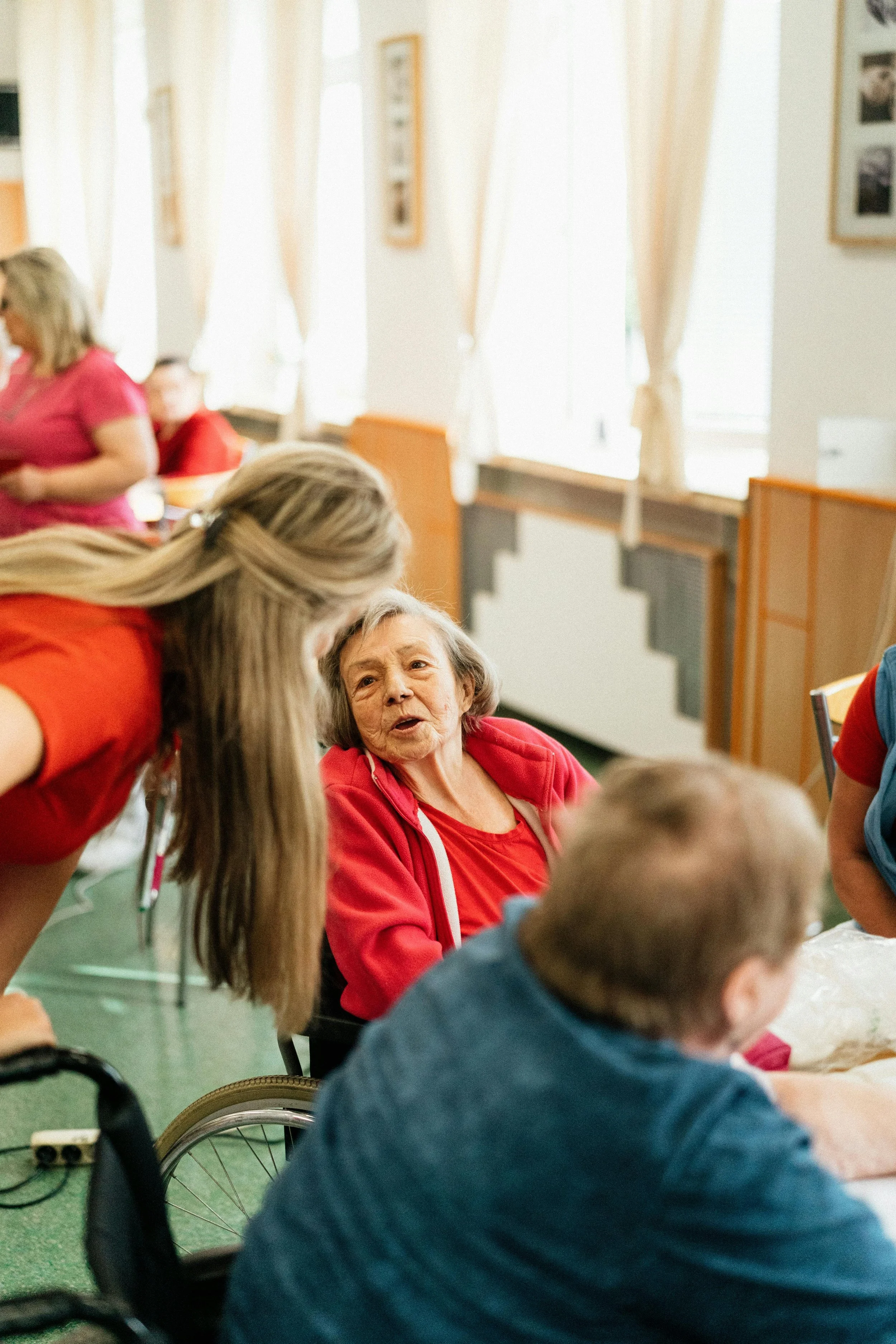 Elderly woman in a red hoodie engaging in conversation with a young caregiver with long blonde hair, in a room with natural light, white walls, and framed pictures.