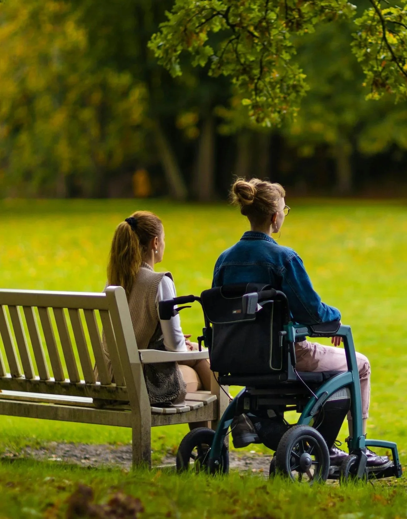 Two women sitting on a park bench, one in a wheelchair, watching the trees and greenery.