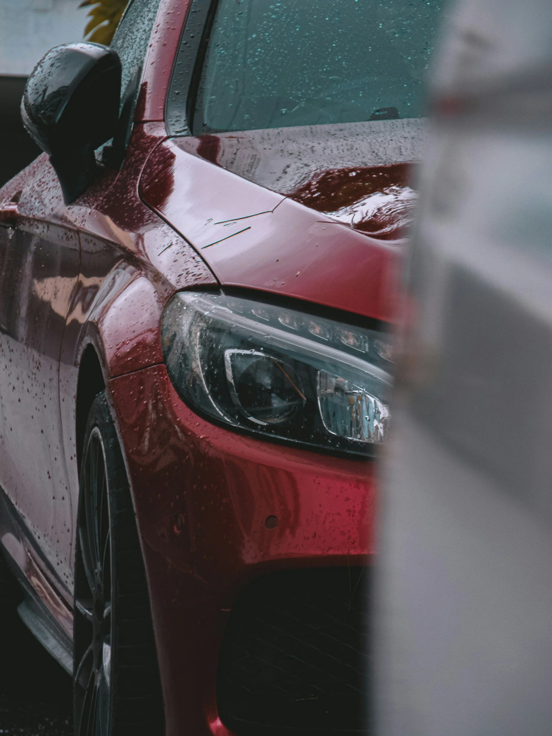 Close-up of a red car's front left side, with rain droplets on the windshield and body, taken during rain.