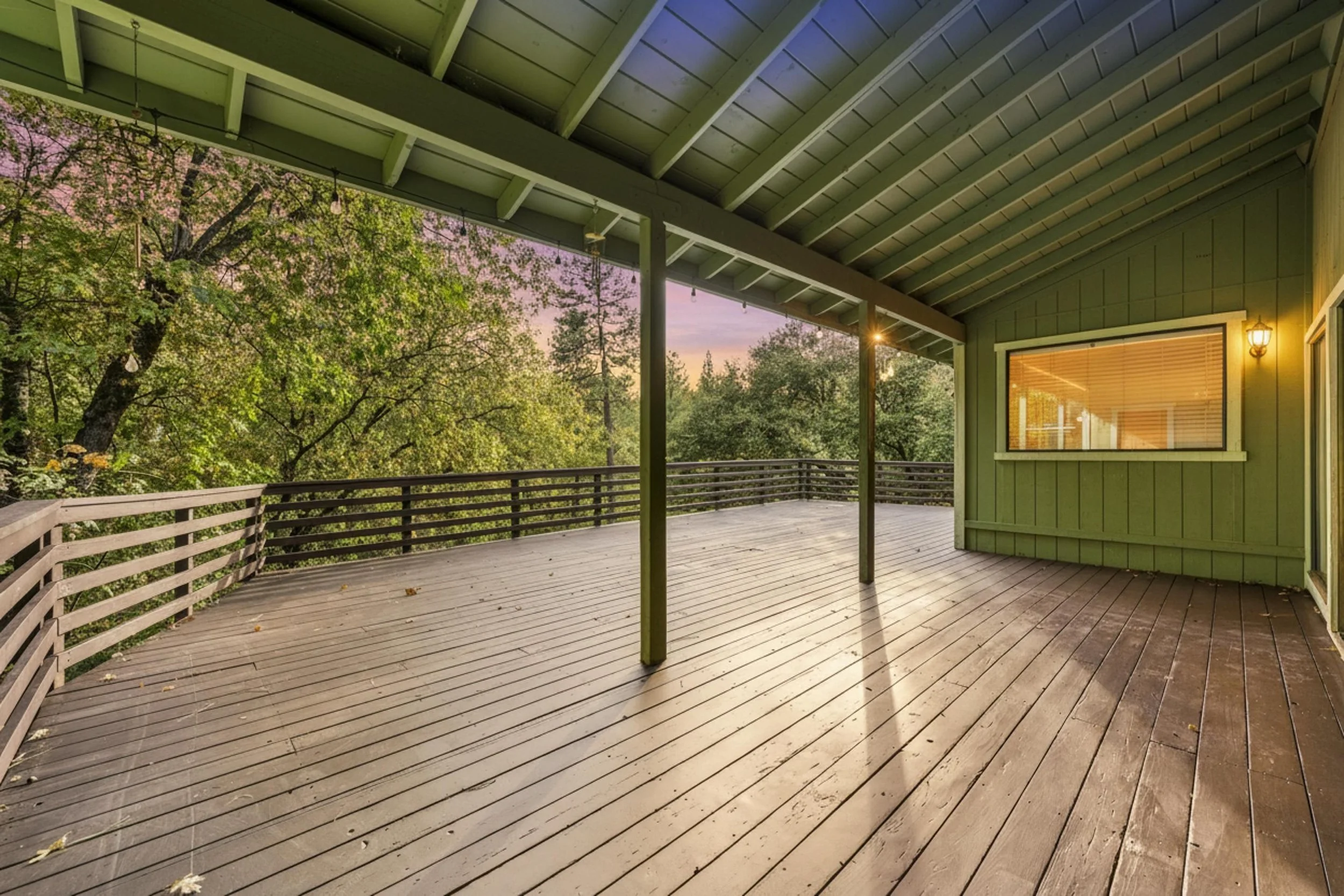 Empty wooden deck with a railing, green exterior house wall with window and outdoor light, overhanging roof, trees with pink blossoms in the background at sunset.