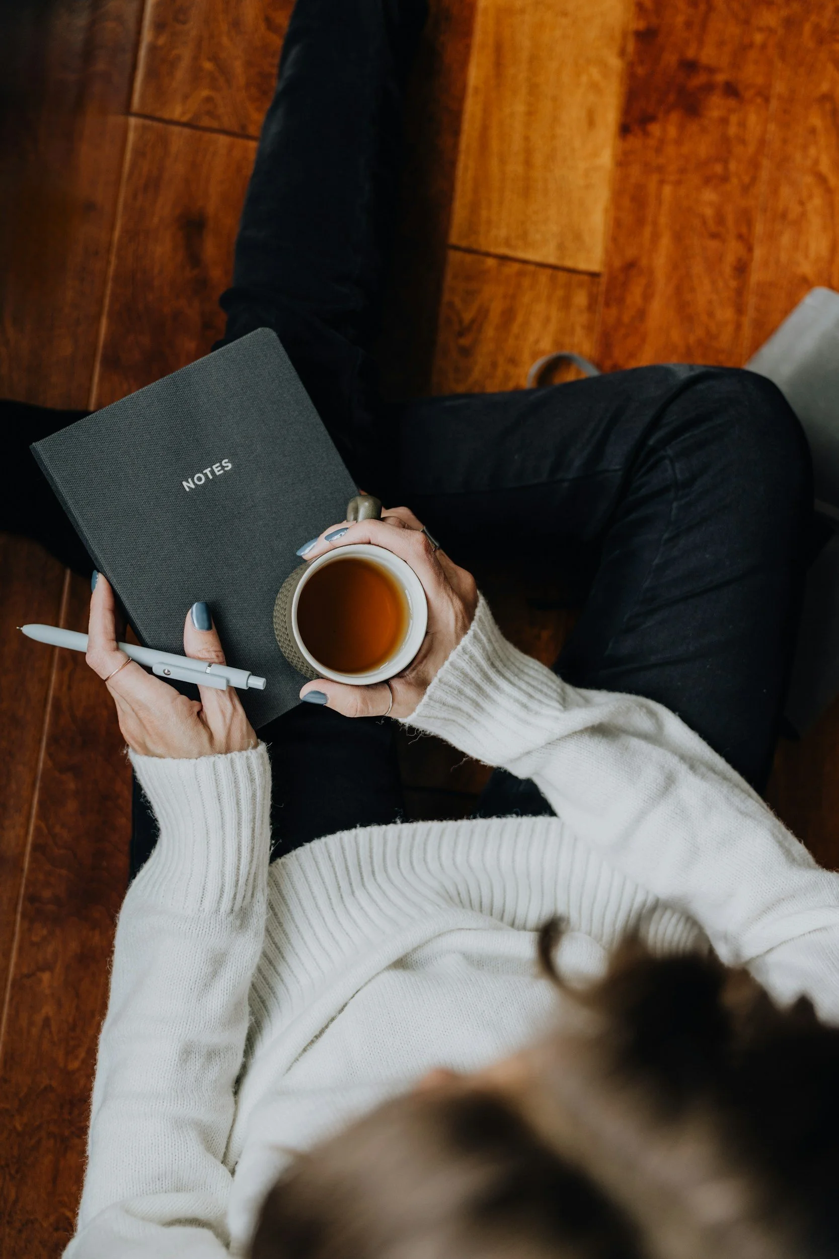Top view of a person wearing a cream-colored sweater holding a mug of tea or coffee, with a notebook, pen, and a person's hand visible, sitting on a wooden floor.