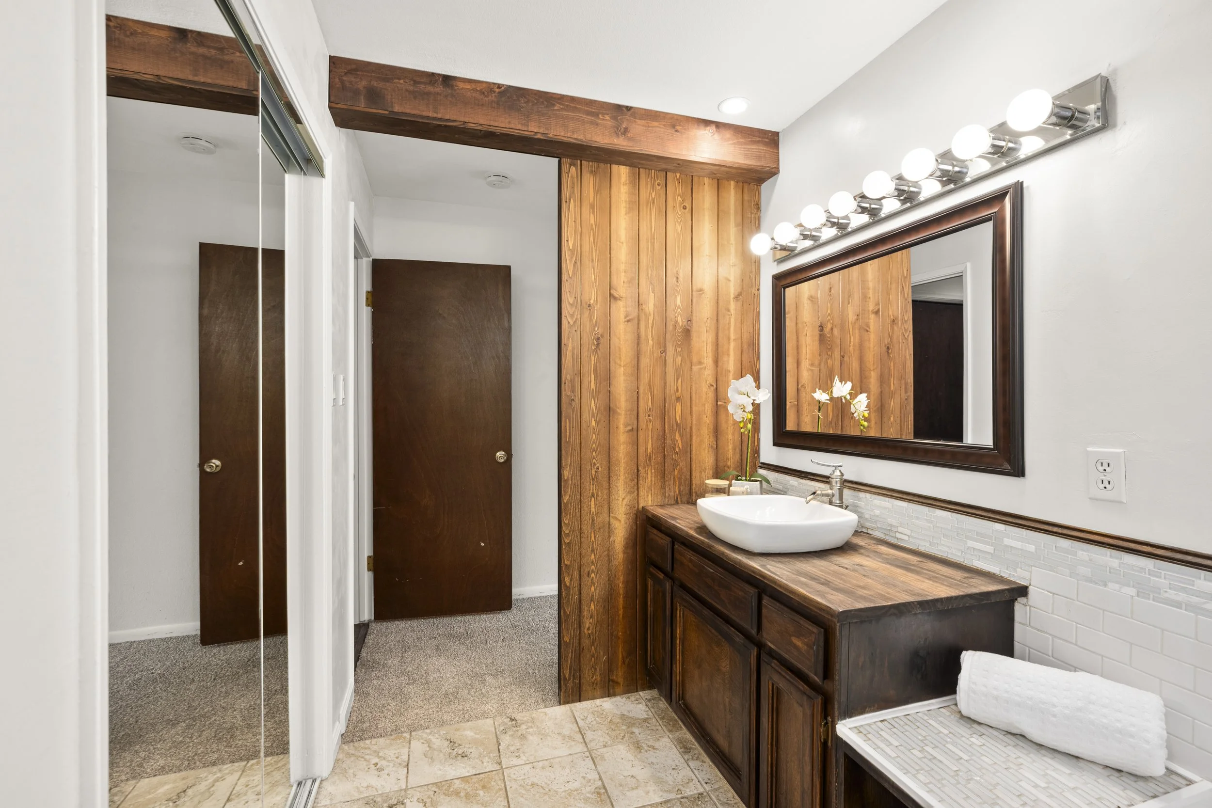 Bathroom with a wooden vanity, a white vessel sink, a large mirror with lights, and wood-paneled wall.