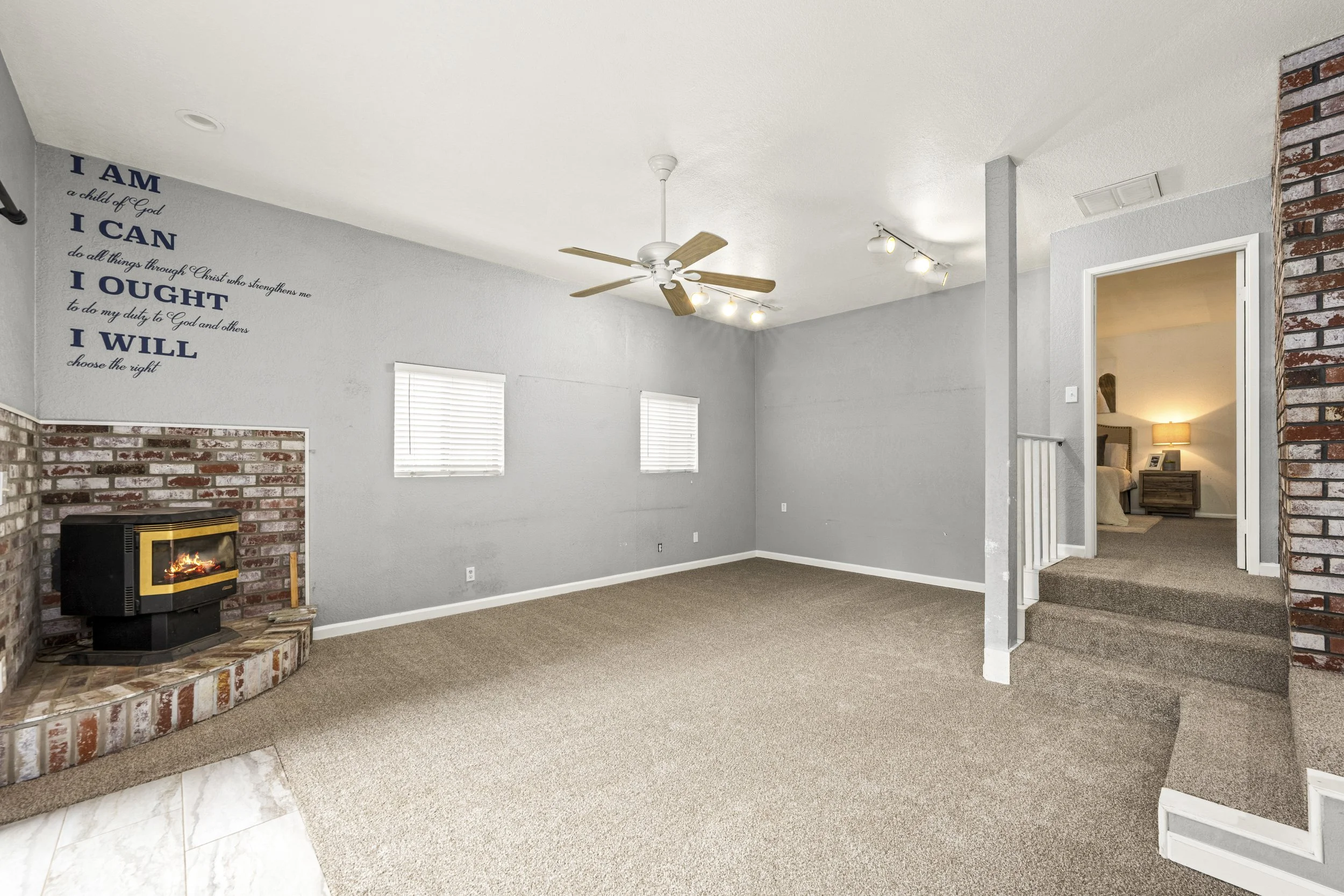 Living room with gray walls, a fireplace with a brick surround, a ceiling fan, two small windows with blinds, and an open doorway leading to a bedroom with a nightstand and lamp.