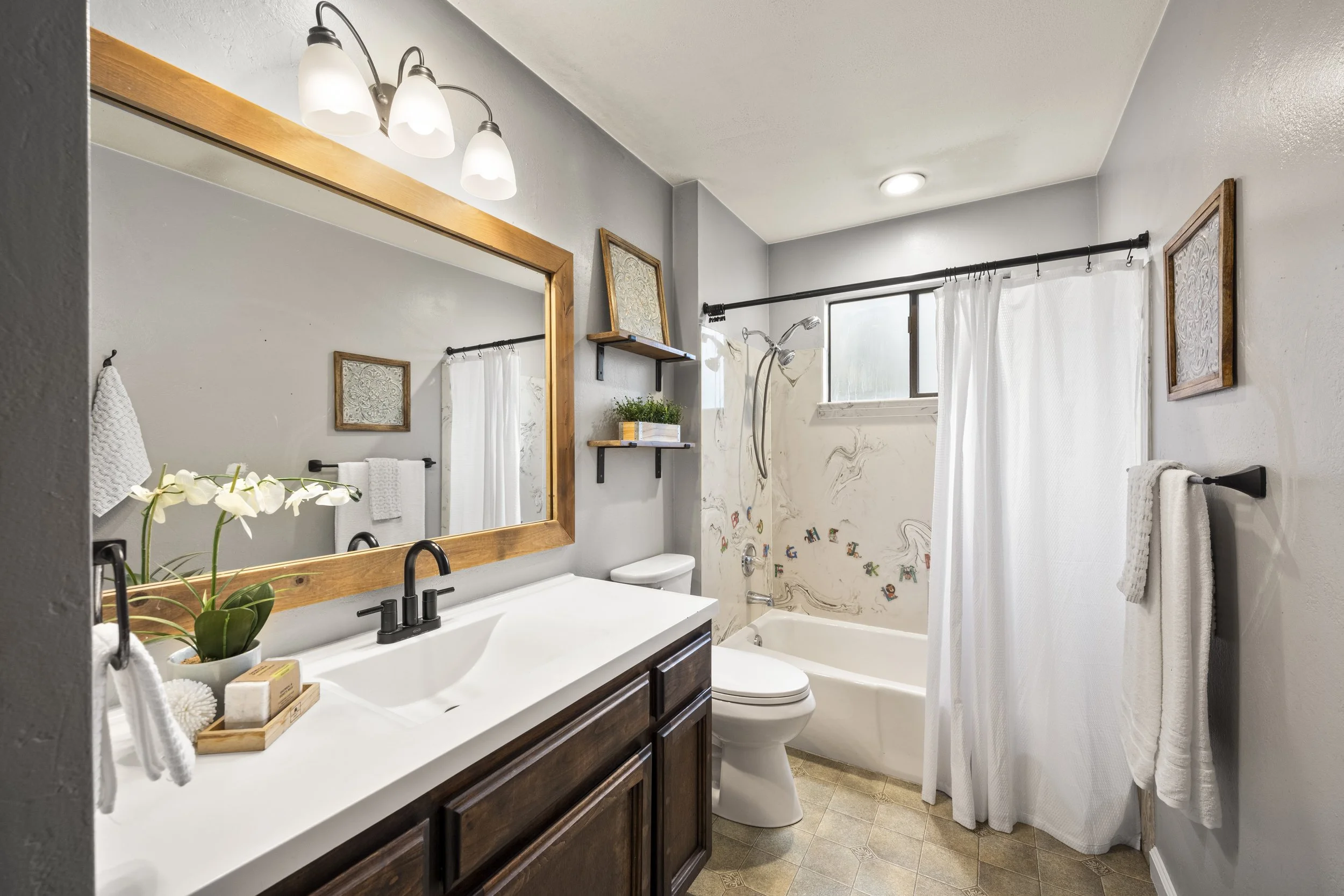 A bathroom with a white countertop sink, wooden vanity, and a mirror framed in wood. There are white towels, a potted plant, and bathroom items on a tray on the counter. The shower area has a curtain, decorative wallpaper, and shelves with books. The