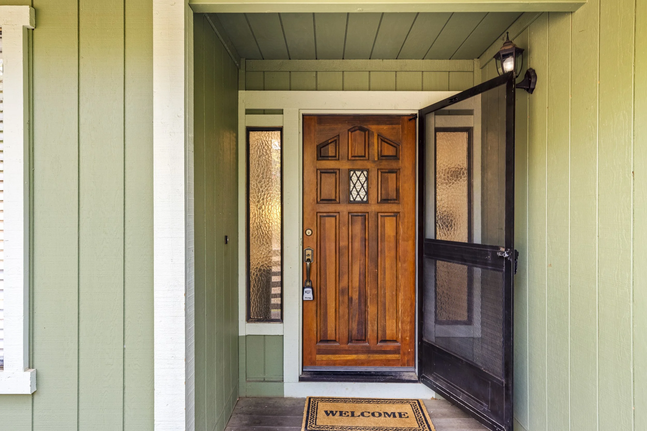 Front door entrance with green siding, a wooden door, and a welcome mat on the porch.