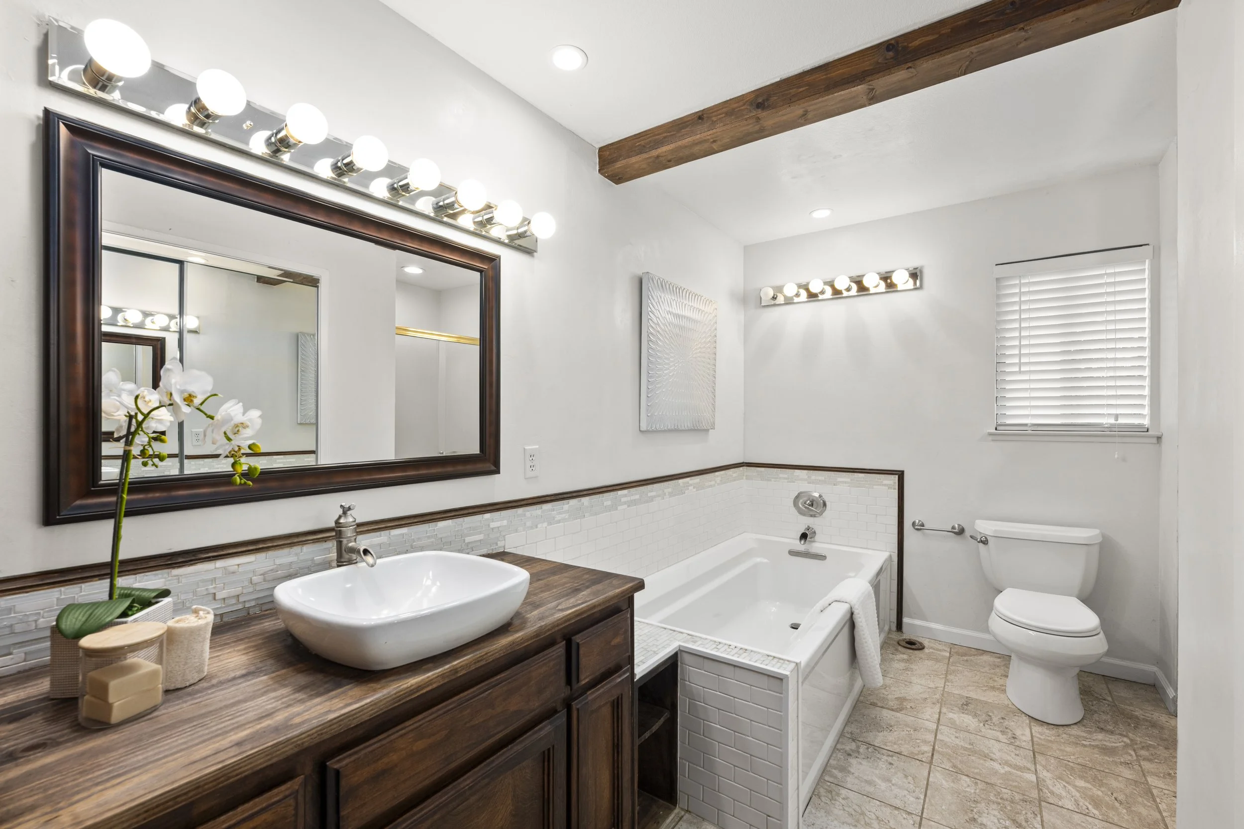 Bathroom with white walls, wooden beams, a large mirror, vanity with a white vessel sink, decorative flowers, and a bathtub with a tile surround and a window with blinds.