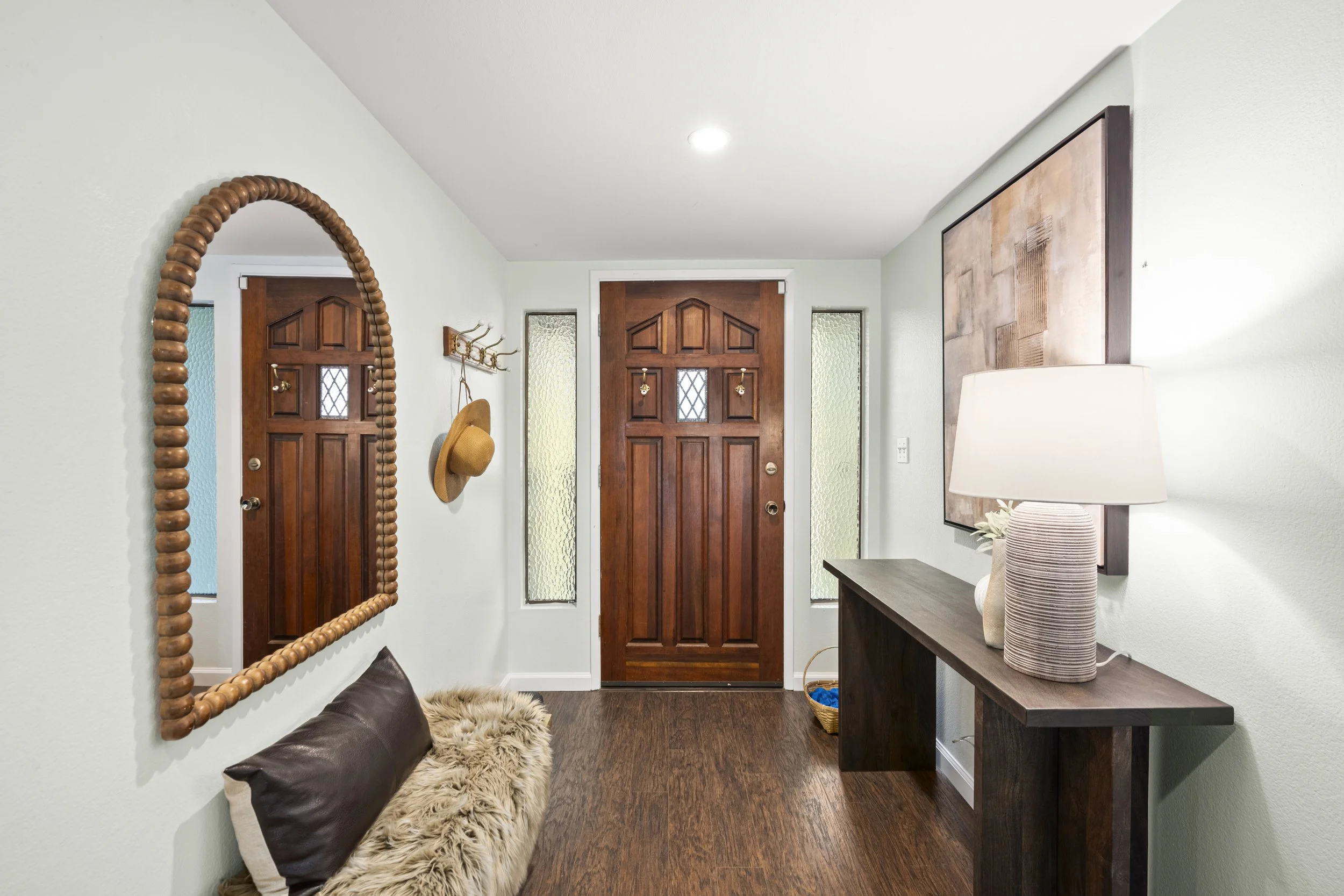 Entryway of a house with a wooden door, a large mirror with a beaded frame, a coat rack with hats, and a console table with lamp and decorative pillow.