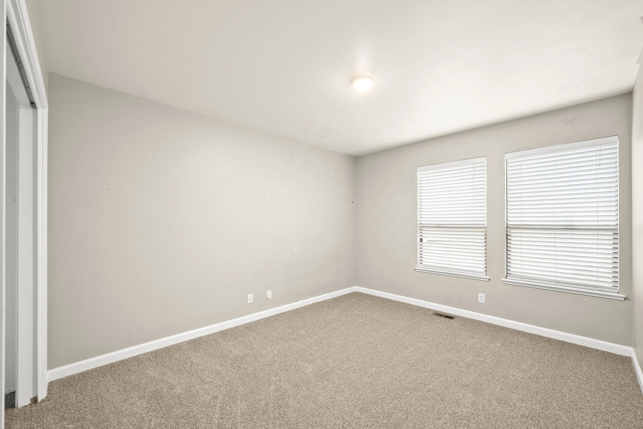 Empty room with beige carpet, white walls, two windows with white blinds, white baseboards, and a ceiling light fixture.