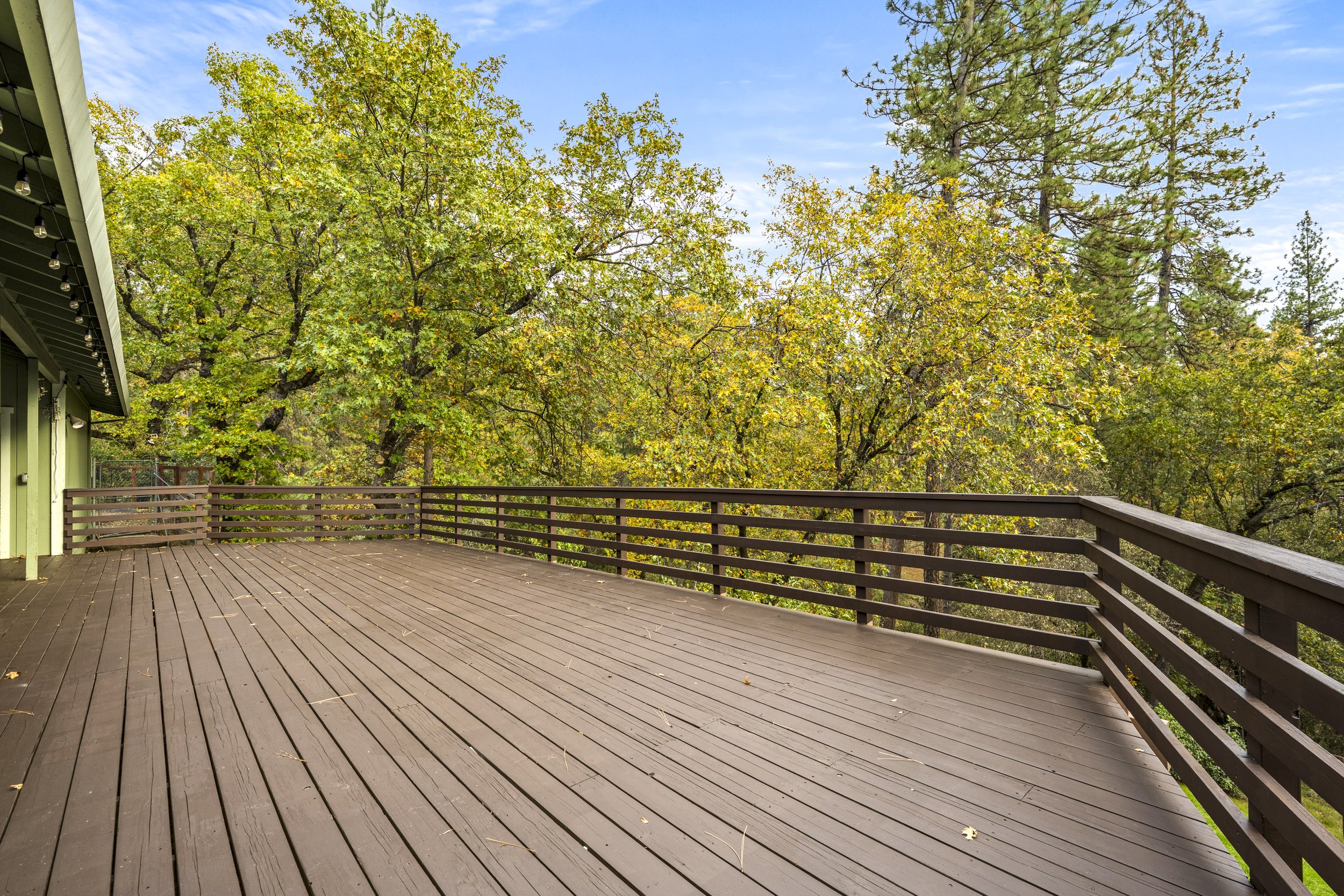 An outdoor wooden deck surrounded by trees with green and yellow leaves, under a partly cloudy blue sky, with a part of a green house or building visible on the left.