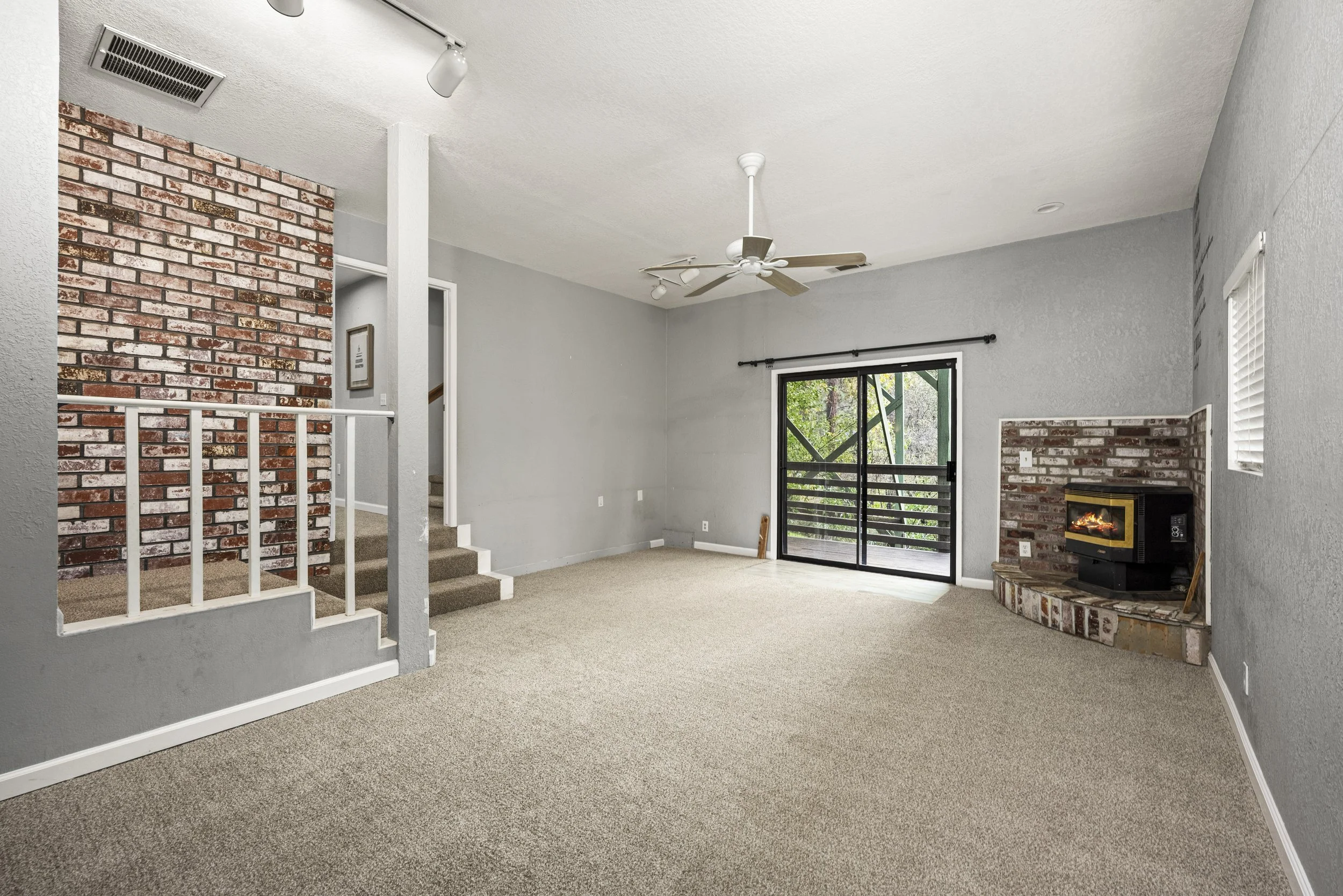 Living room with beige carpet, gray walls, brick fireplace, ceiling fan, sliding door leading to outdoor space, and window with blinds.